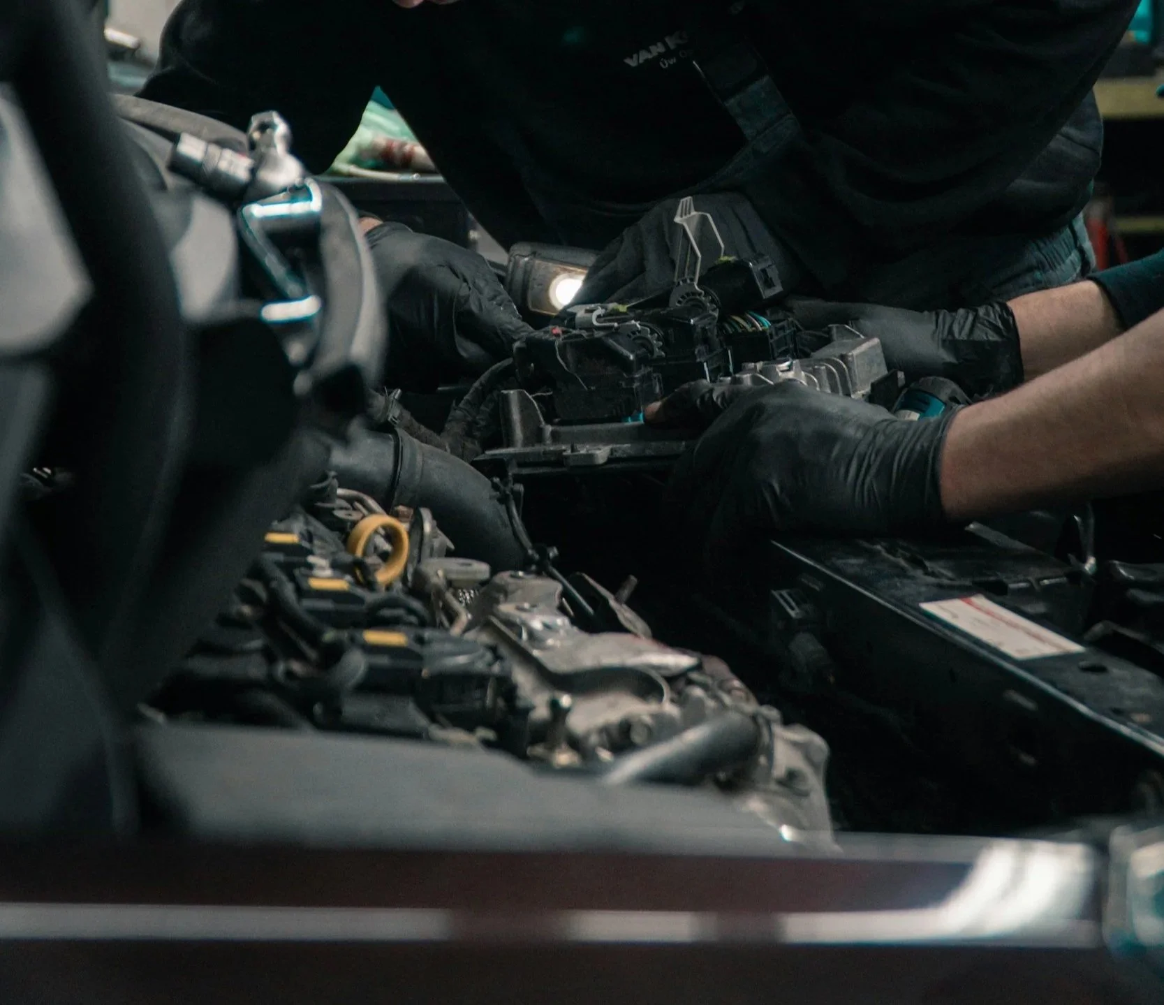 A mechanic working on a car engine with a flashlight in hand, wearing gloves.