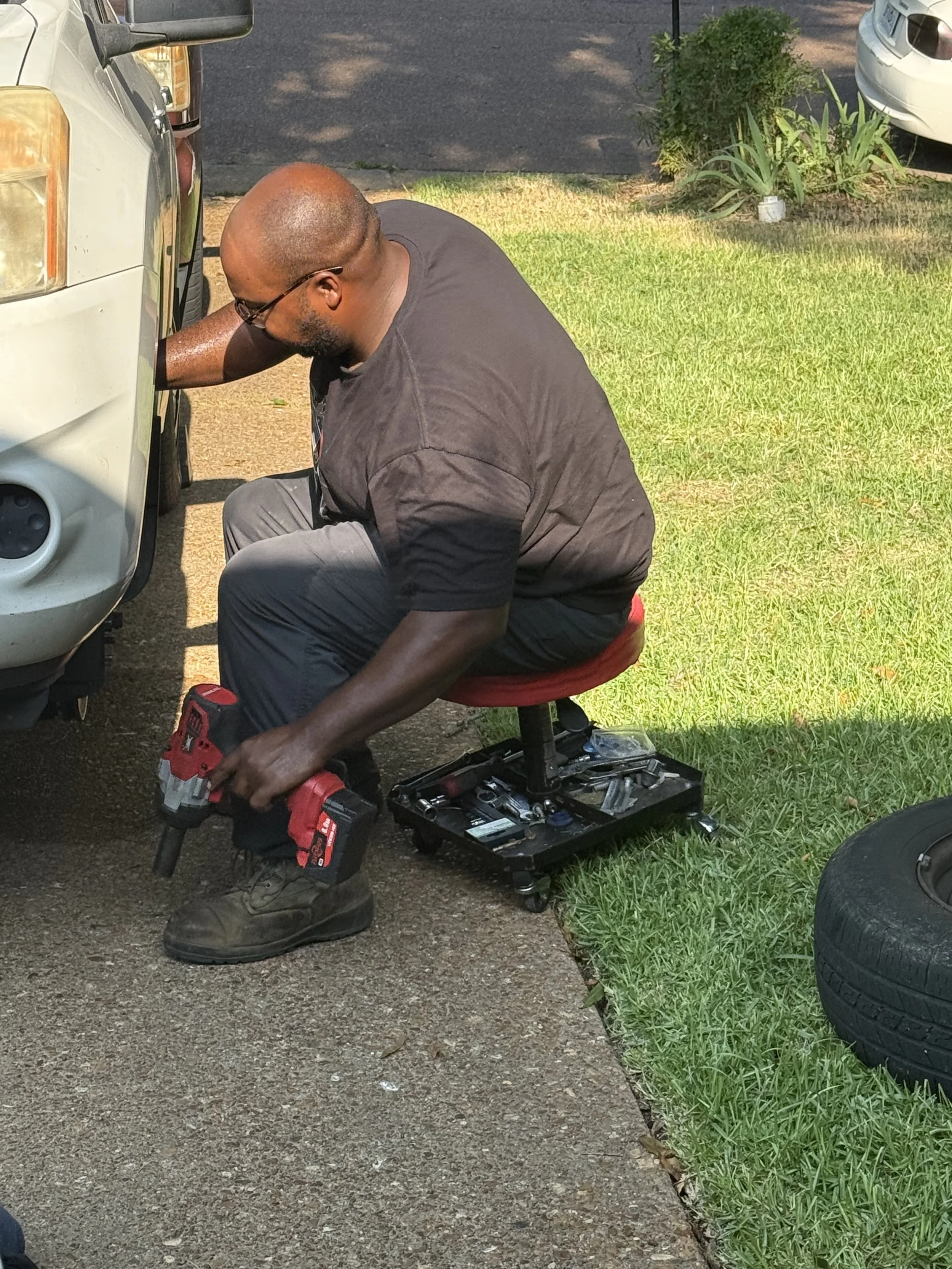 Man working on a vehicle's front wheel with tools, sitting on a mechanic's stool outdoors near cars and plants.