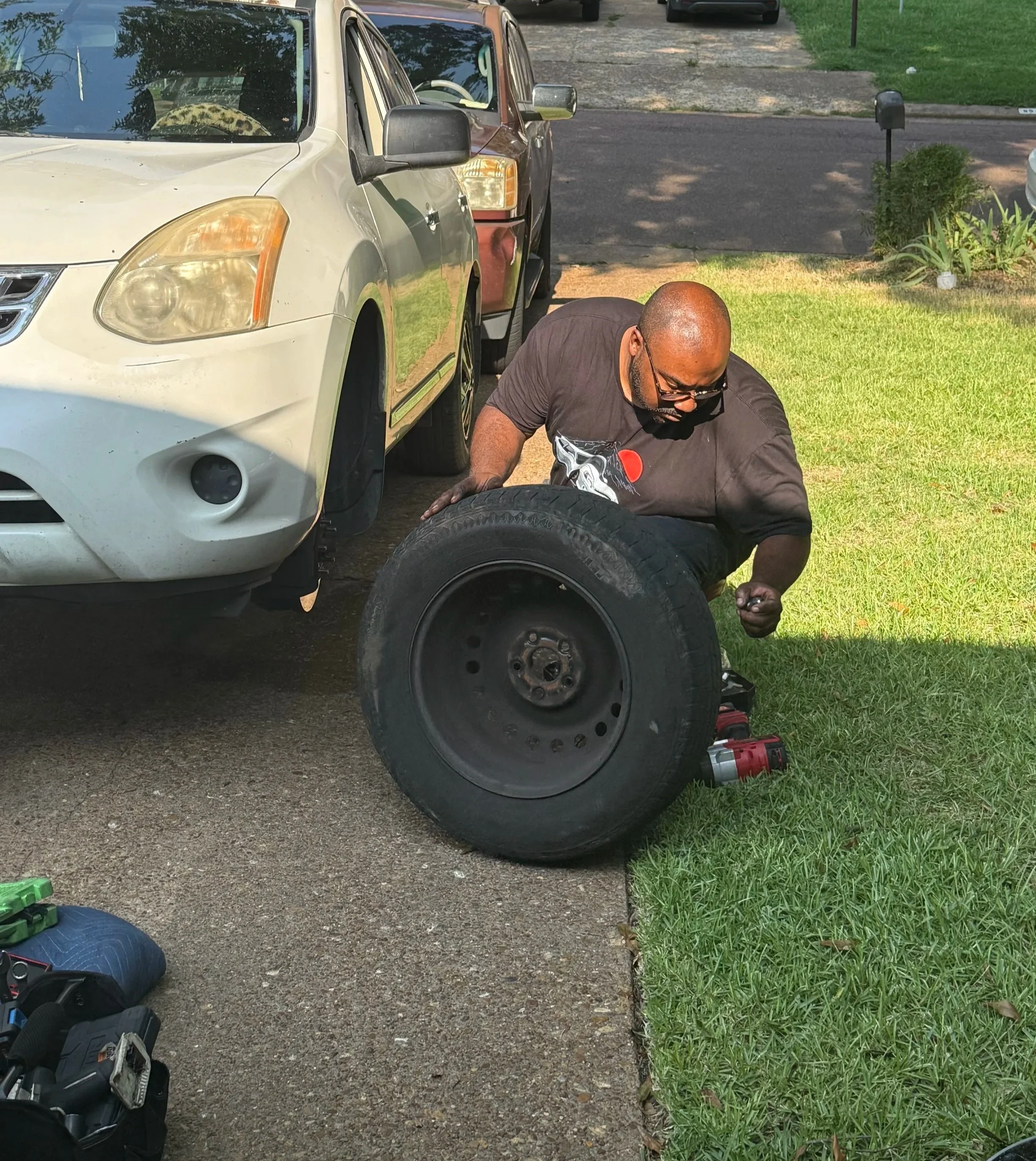 A man working on a tire alongside a white and maroon car parked on a driveway. Tools and equipment are nearby, and there is grass and greenery on the side of the driveway.