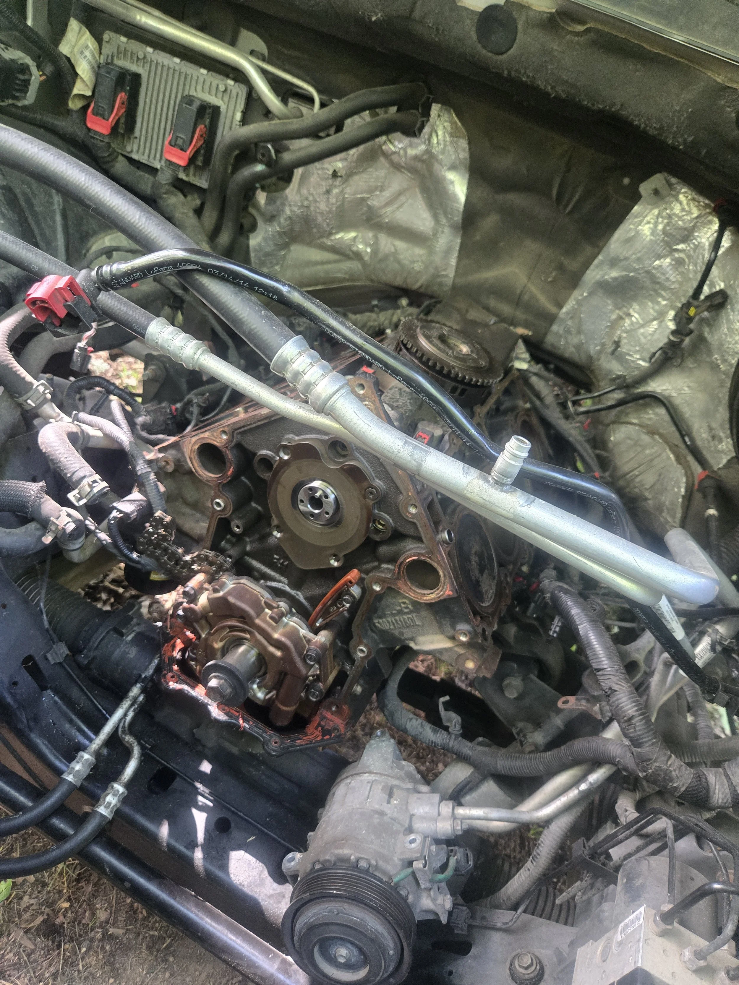 A partially disassembled car engine showing various belts, pulleys, hoses, and components, with the engine cover removed.