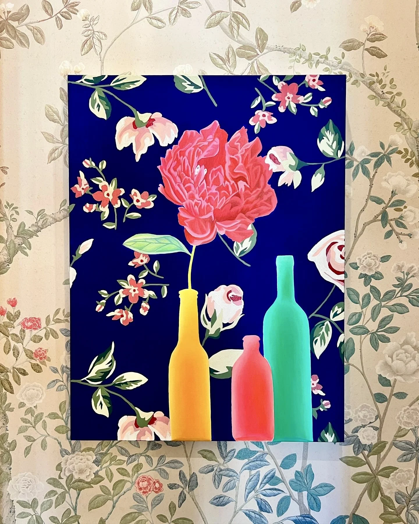 A colorful painting of three bottles in yellow, pink, and teal with a large pink peony and other flowers and leaves on a dark blue background, framed by light floral wallpaper.