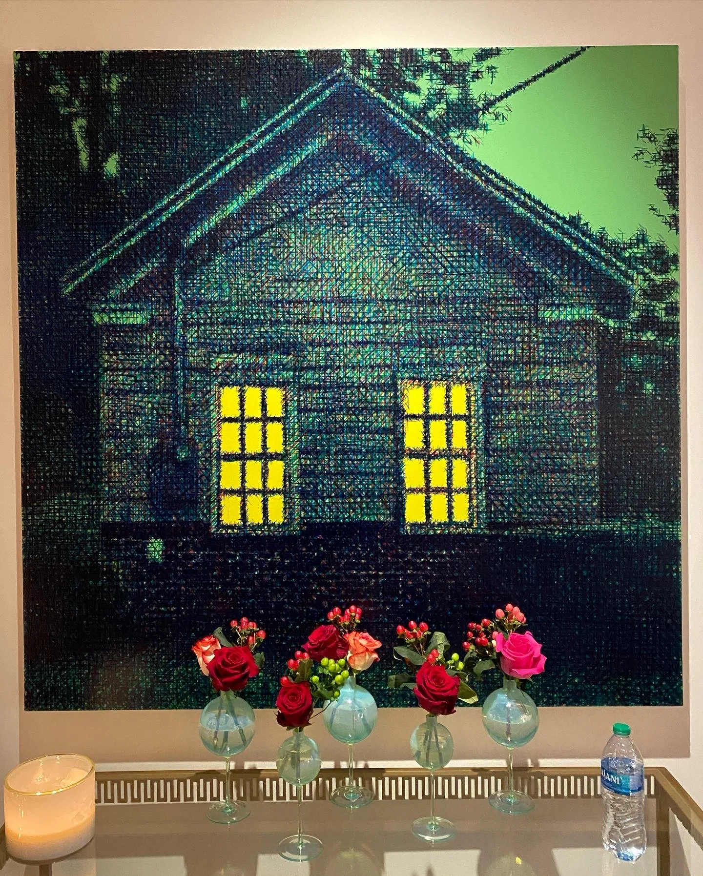 A wall with an illuminated house-shaped artwork, glowing yellow windows, and surrounded by red and pink flowers in vases placed on a table, with a candle and a bottle of water nearby.