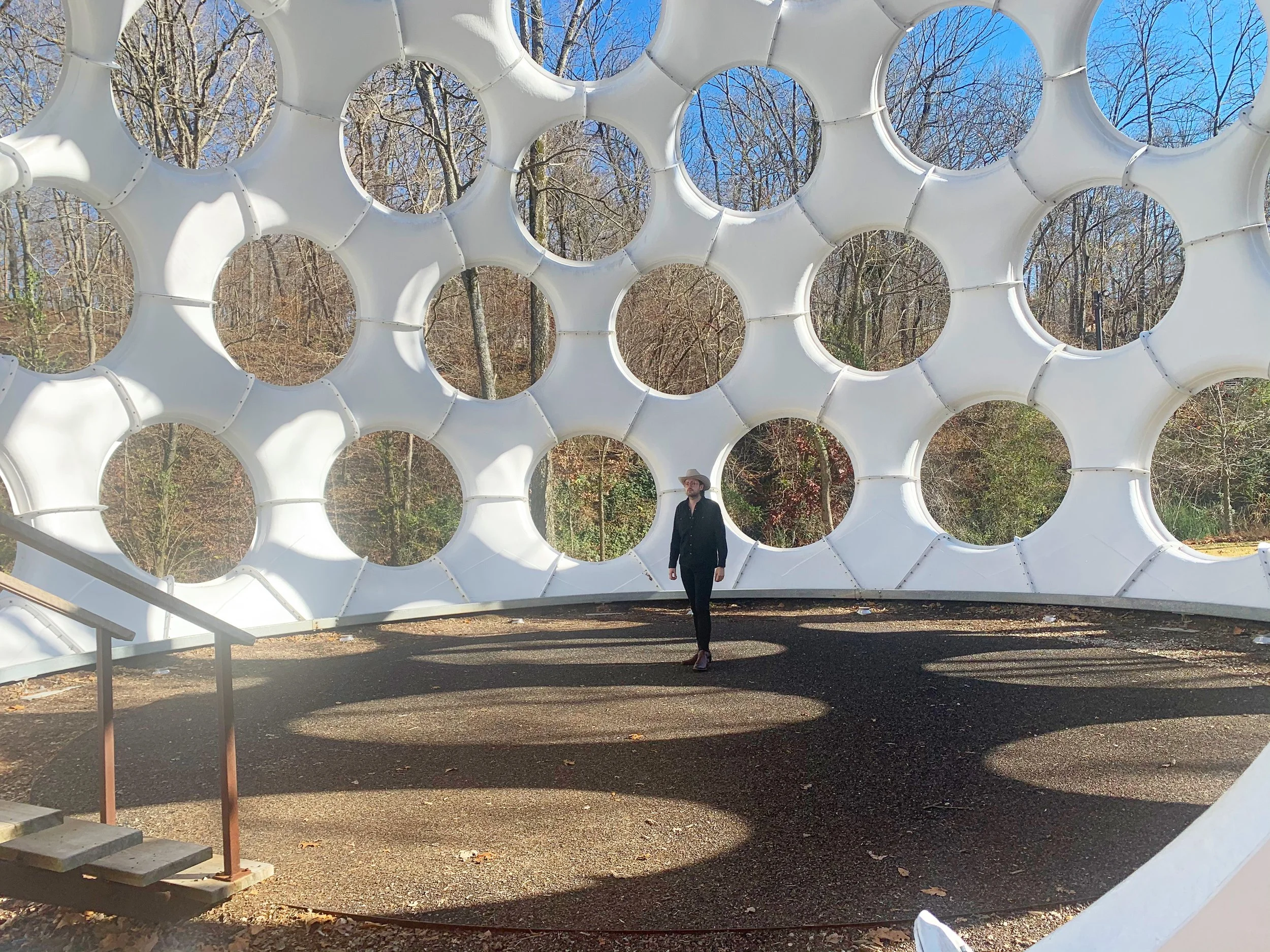 A person wearing a black outfit and a wide-brimmed hat standing under a large, white, circular, petal-like sculpture outdoors during daylight. The background features leafless trees and a bright blue sky.