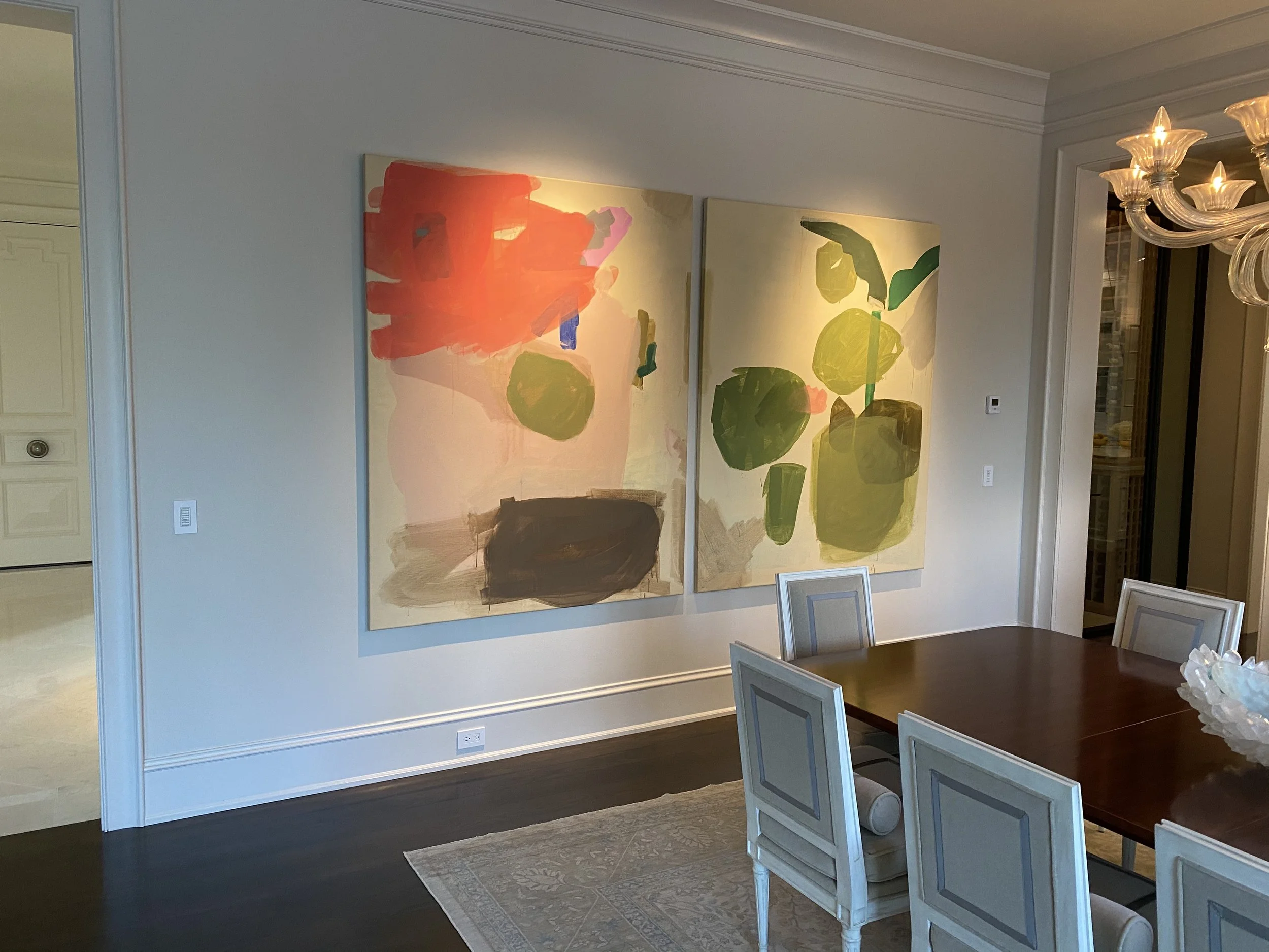 A dining room with two abstract paintings of large green and red forms hanging on the wall, a chandelier with multiple glass arms and warm lighting, and a wooden dining table with light-colored chairs.