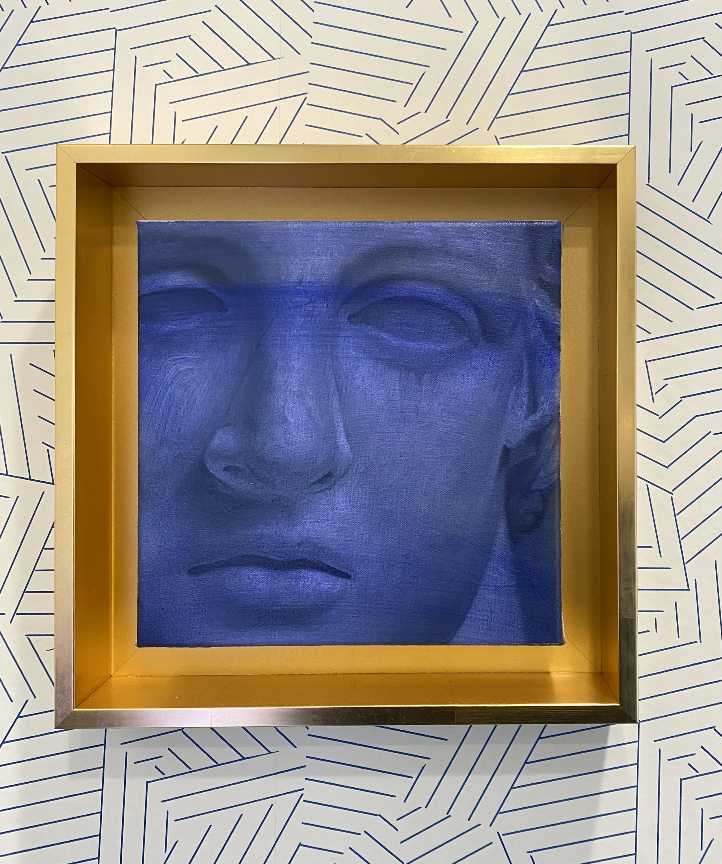 A framed artwork of a face with closed eyes and a neutral expression, displayed on a wall with a blue geometric line pattern background.
