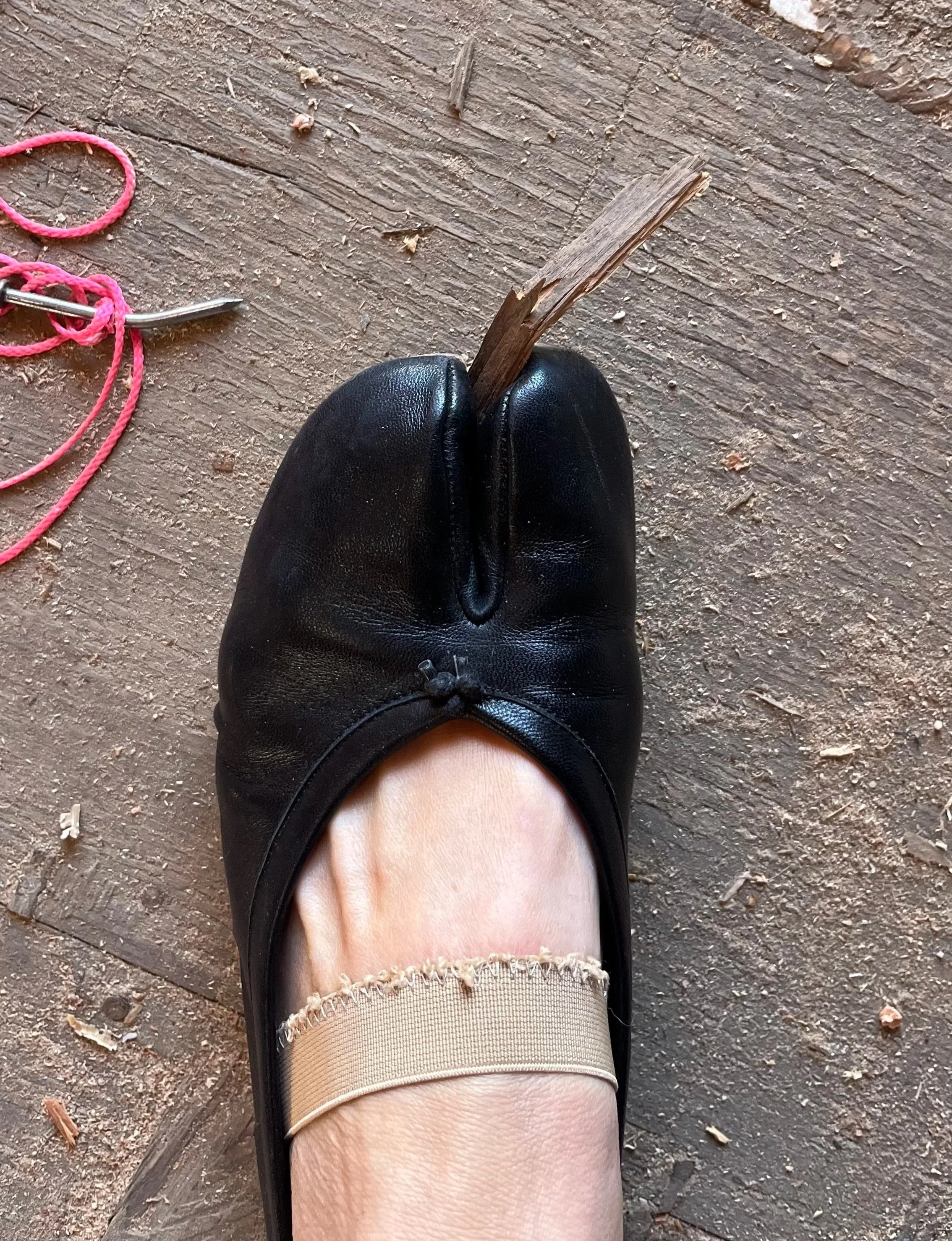 From a top-down perspective, a person is wearing a black shoe on their left foot, with the foot on a wooden surface. A beige sock with lace trim can be seen above the shoe, and a small part of their leg is visible. To the left, a pink shoelace and a metal shoehorn are on the wooden surface. A chipped wooden stick is placed vertically between the person's foot and shoehorn.