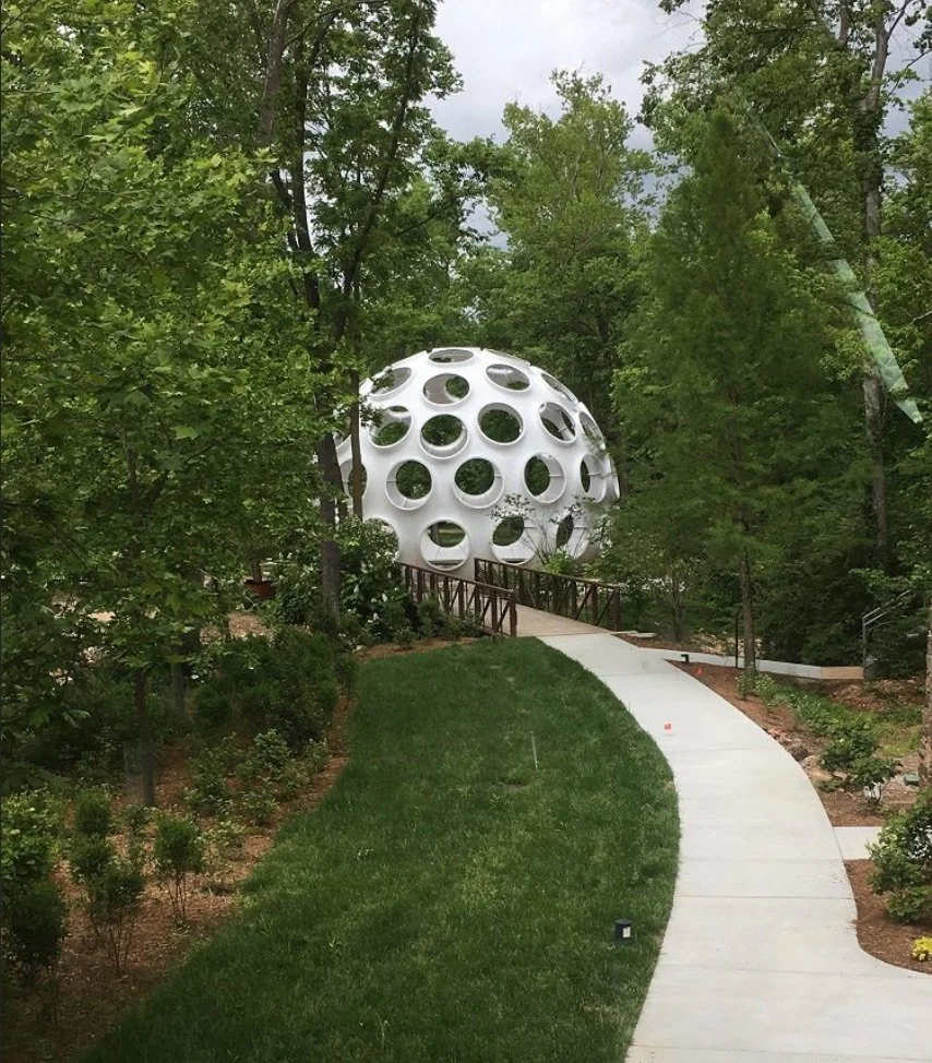 A modern, spherical house with large circular windows on a hillside, surrounded by green trees and a grassy yard, connected to a sidewalk via a small wooden ramp.