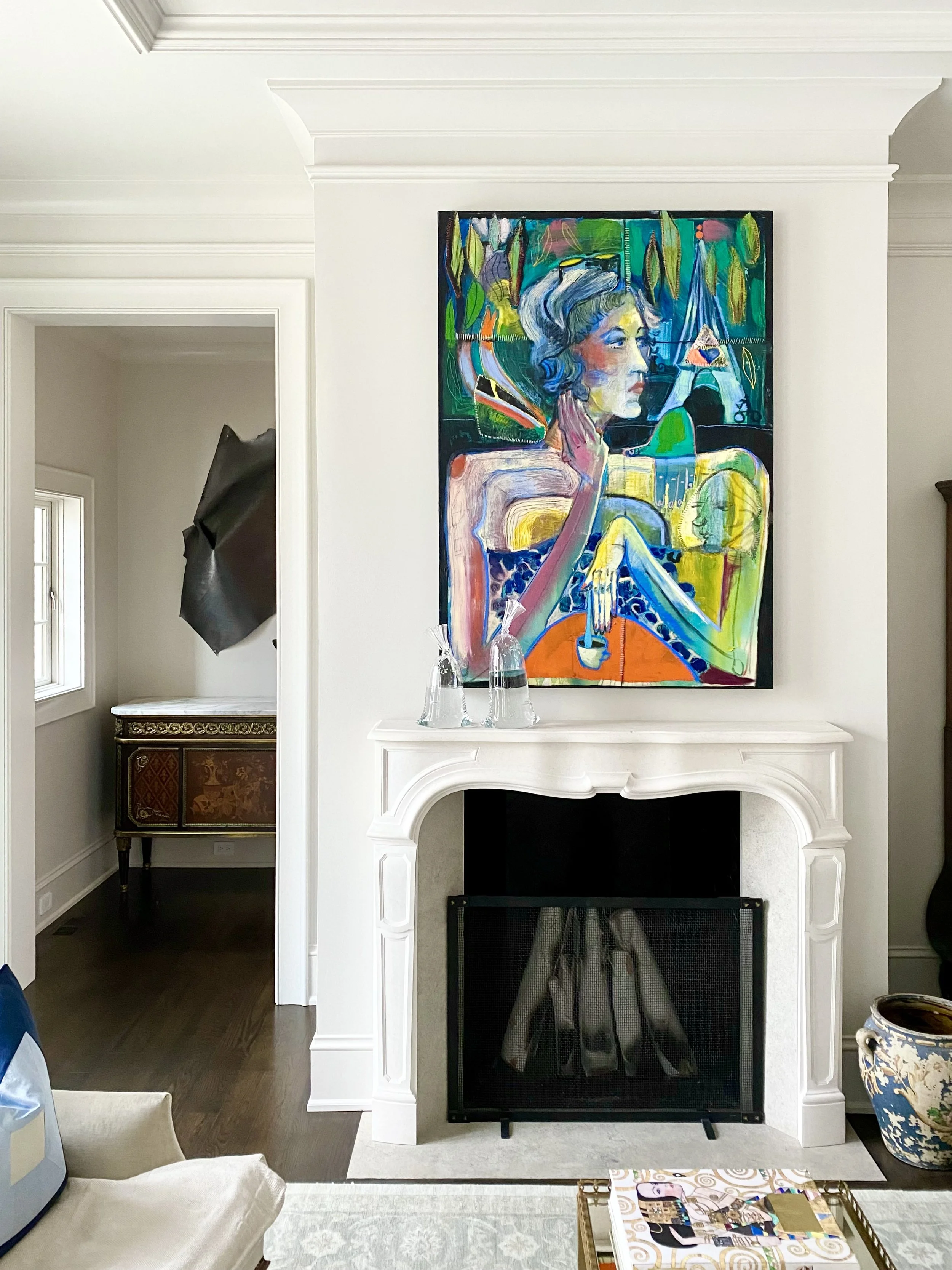 A bold colorful figurative painting by Kate Barbee prominently hangs above the marble carved fireplace in this traditional contemporary home, Dylan Martinez glass water bag sculptures, and a Nancy Rubins wall drawing fill the space.