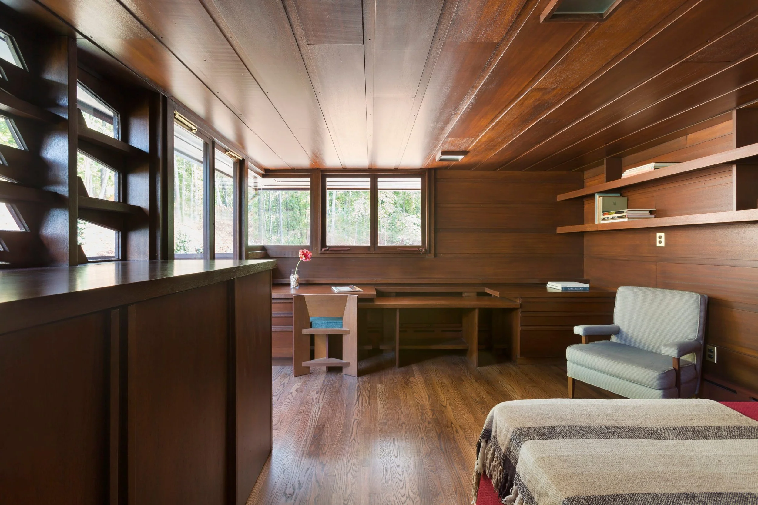 Board and batten mahogany walls and ceiling, wrap the mass of this Frank Lloyd Wright designed bedroom, glass crank casement window, built in desk, and ornamental carved windows easily identify the characteristics of Frank Lloyd Wright.
