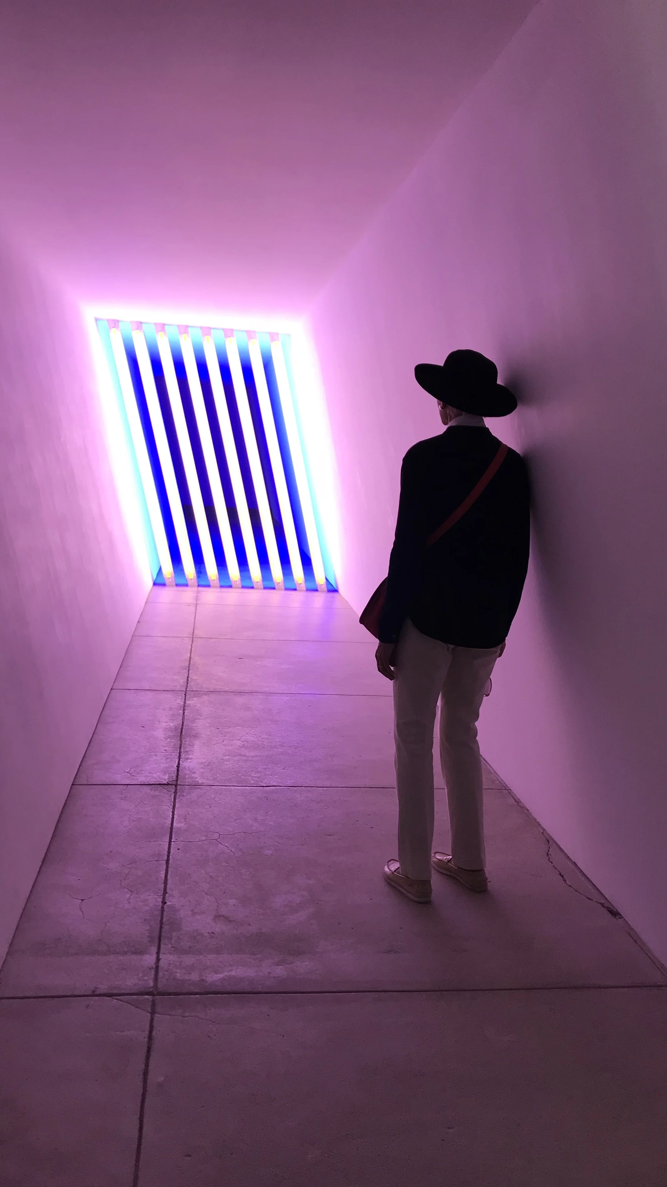 A person with a large black hat and beige pants standing in an indoor space with pink walls, looking at a colorful light installation featuring vertical blue and yellow stripes, illuminated with pink and purple lighting.