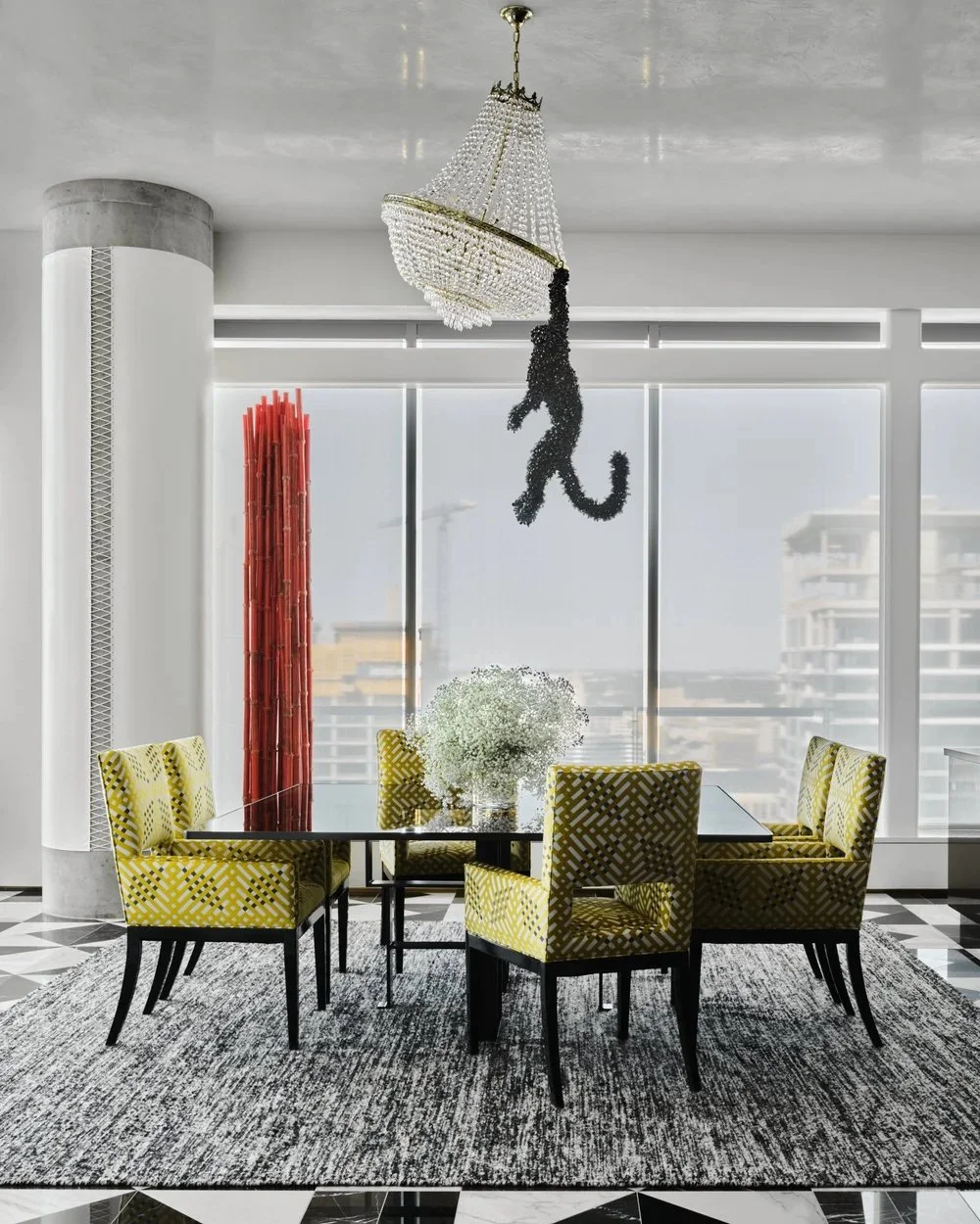 Modern dining room with a black table, yellow patterned chairs, a gray area rug, large windows, a chandelier, and a decorative hanging sculpture.