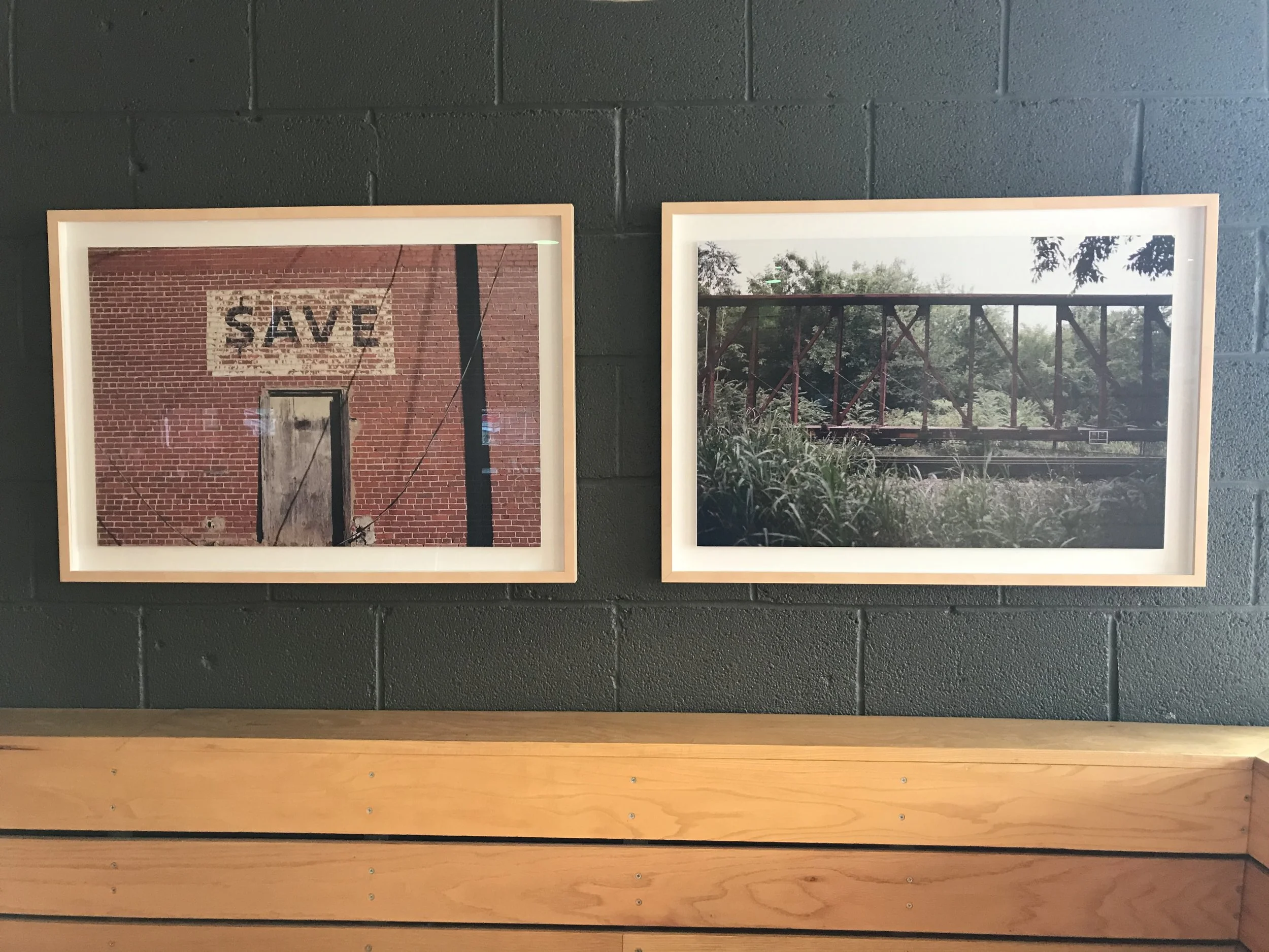 Two framed photographs hanging on a black brick wall. The left photo shows a brick wall with a sign that says 'SAVE,' underneath a boarded-up door. The right photo shows a bridge over a river, surrounded by greenery.