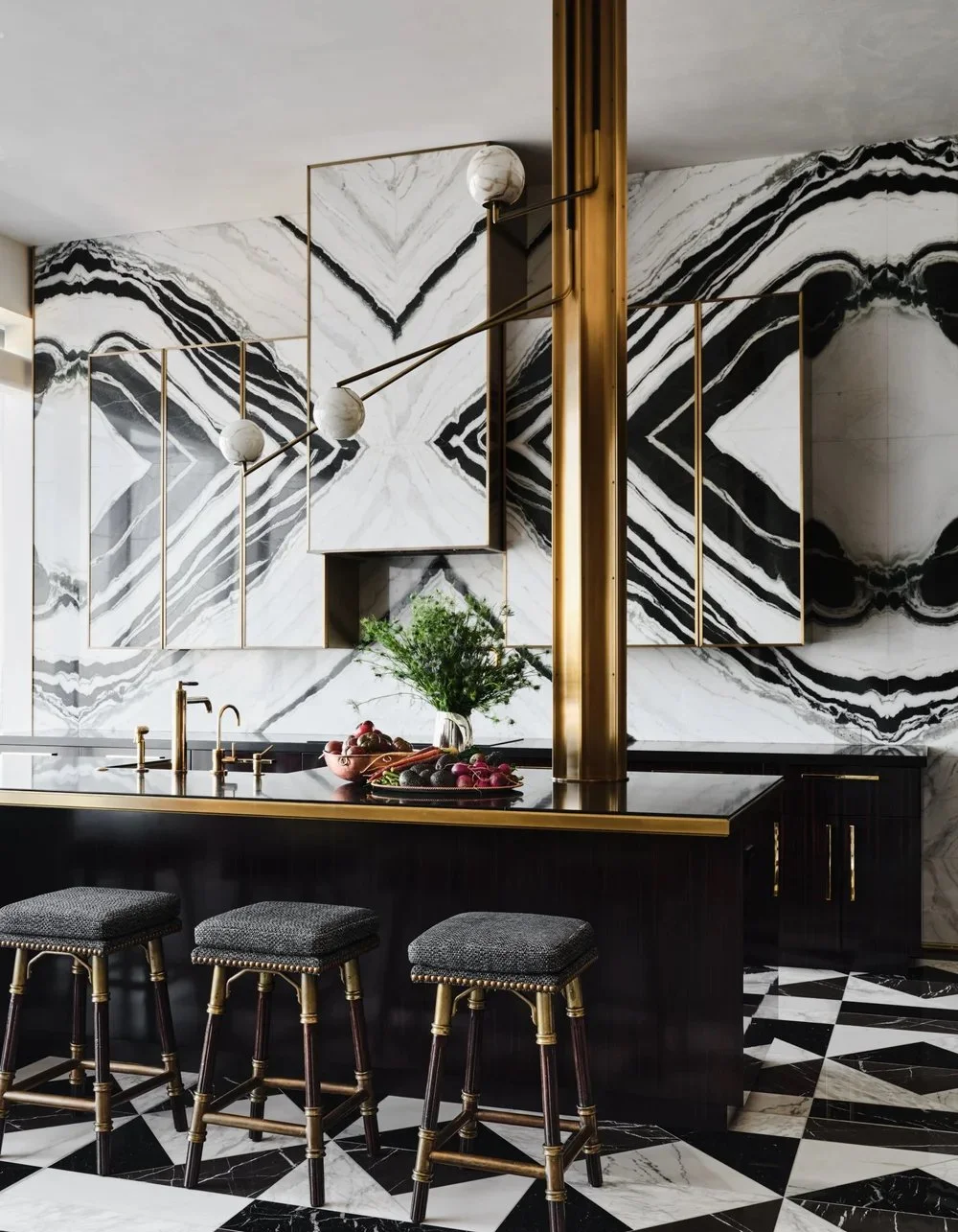 Luxury kitchen with black and white marble walls and floors, gold accents, a black island with gold trim, three black bar stools with gold legs, a green plant, and fresh vegetables on the counter.