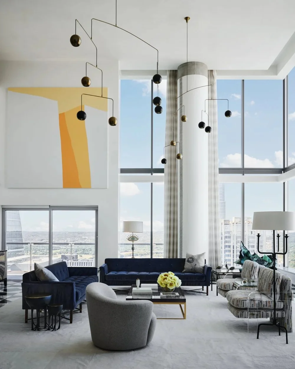 Modern living room with floor-to-ceiling windows, a blue velvet sofa, a light gray armchair, and a patterned sofa. A glass coffee table with a flower arrangement is in the center. There are abstract paintings on the wall and a contemporary chandelier hanging from the ceiling. Large curtains frame the windows, revealing a city skyline.
