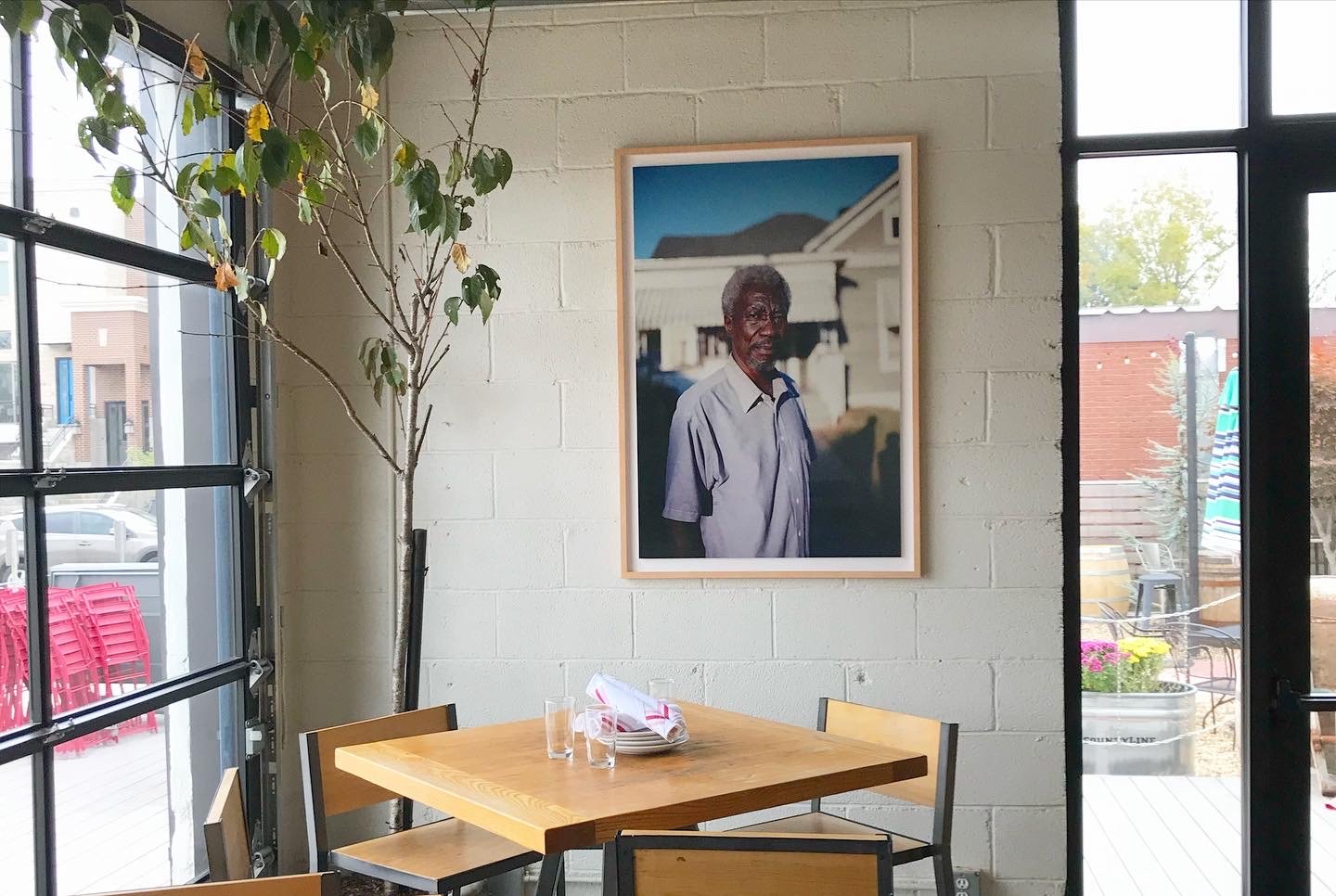 Interior view of a restaurant with a wooden table set with napkins and glasses, a potted plant, and a framed portrait of an elderly man on a white brick wall, with large windows showing outdoor seating and buildings.