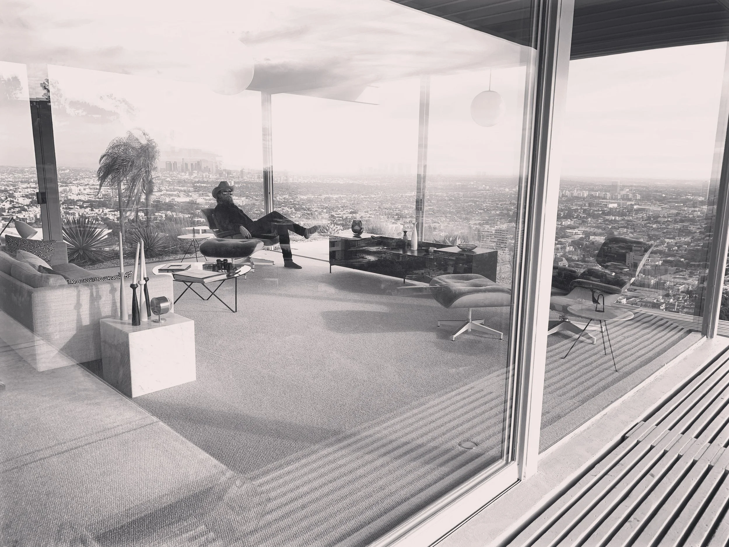 A modern living room with large glass windows overlooking a cityscape. Inside, there is a man wearing sunglasses and a hat, sitting relaxed on a lounge chair with his legs crossed. The room has minimalistic furniture, including a low coffee table, and decorative items like candles and vases. There are plants outside and the space is well-lit with natural light.