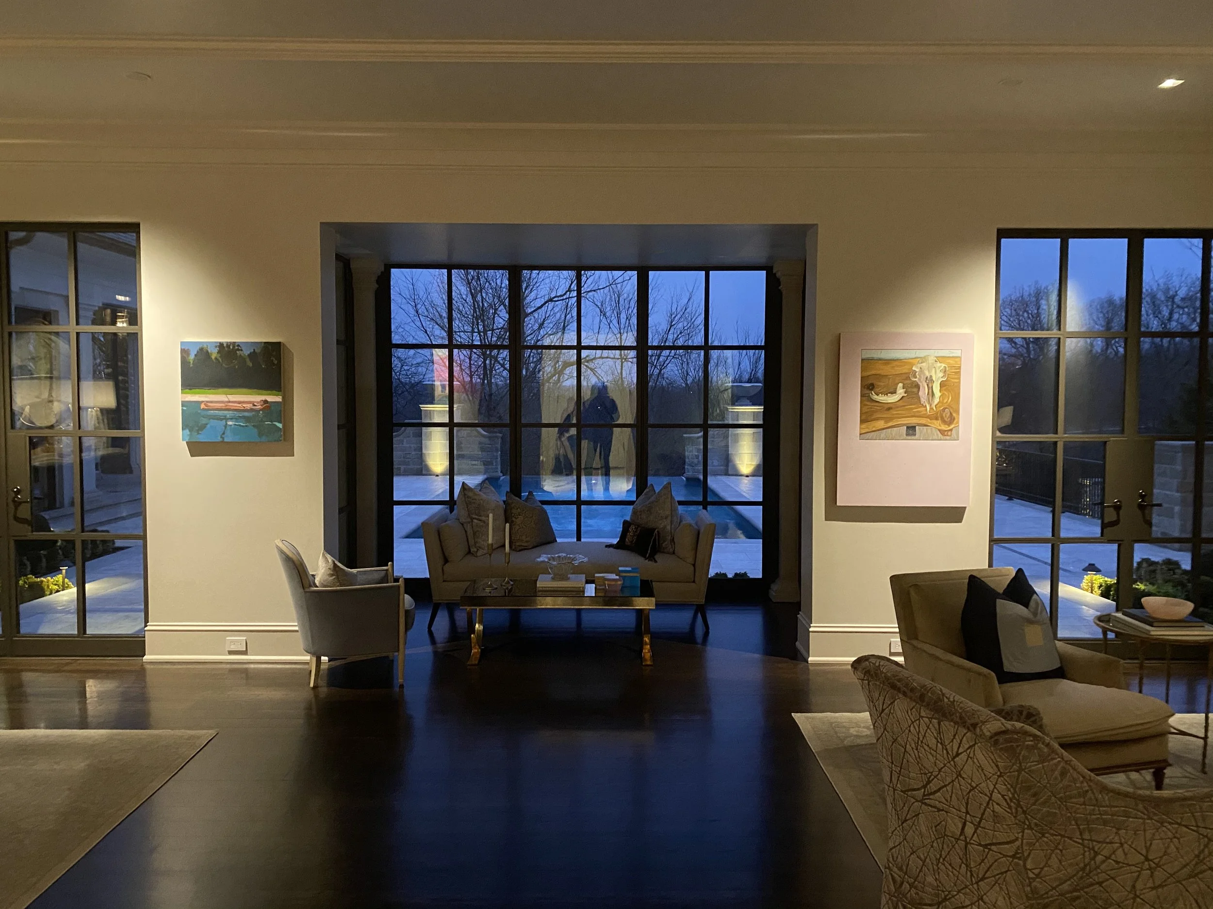 Living room with large windows and glass door, showing a view of trees outside at dusk. Artworks hang on the walls, and there are various chairs and a sofa inside.