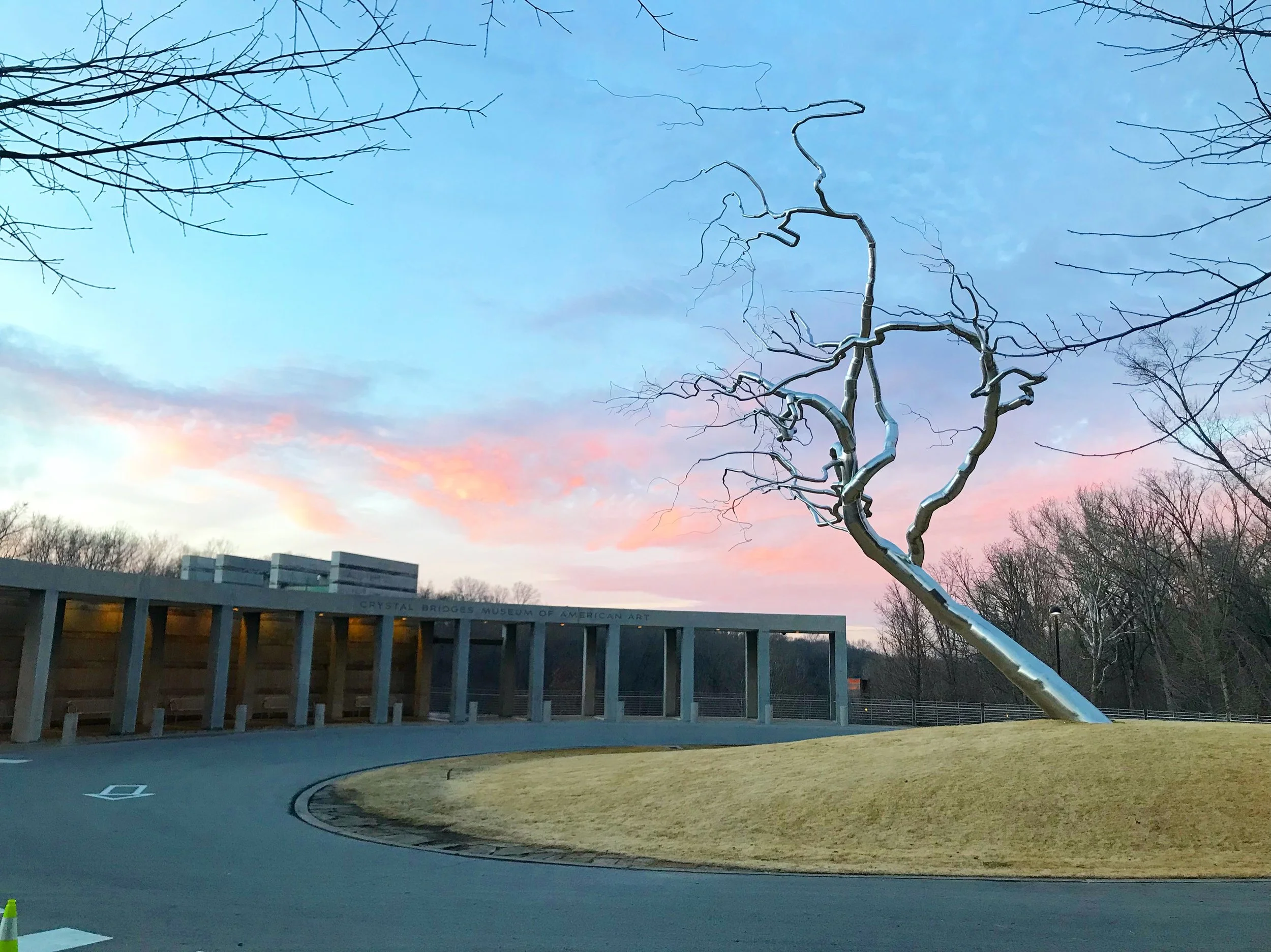A modern sculpture of a leafless tree with metallic, twisted branches in front of a museum building with a sign that reads 'Crystal Bridges Museum of American Art.' The sky is at sunset with pink and blue hues, and there are leafless trees surrounding the scene.