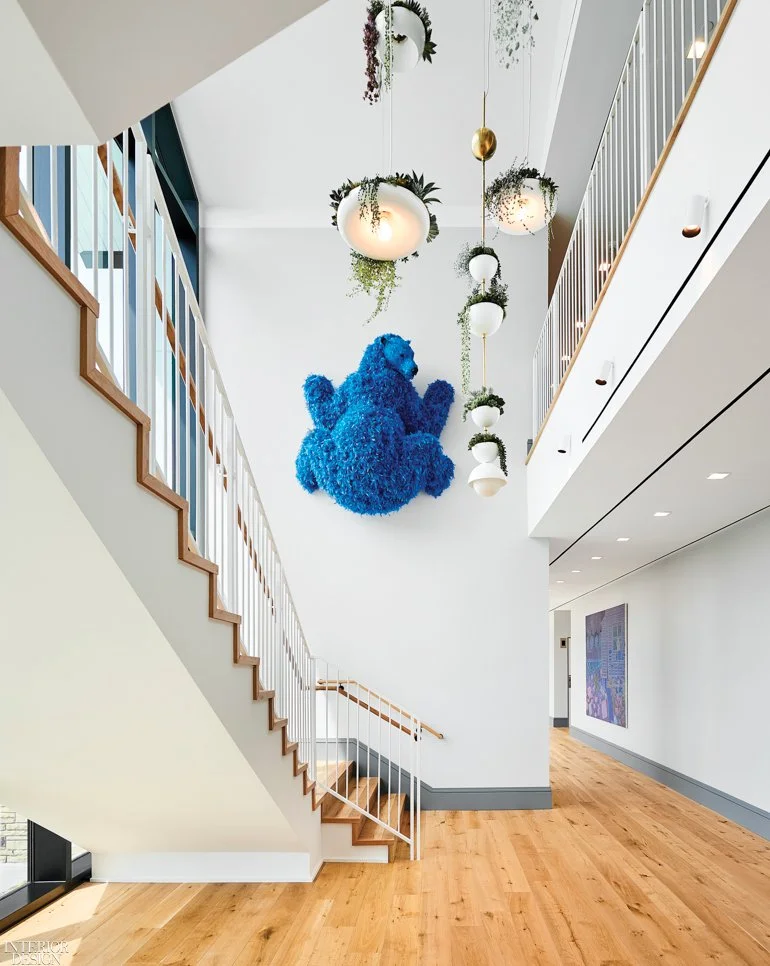 Contemporary stairwell, light wood floors, a blue feathered bear sculpture hangs on the wall above the room, hanging light fixtures filled with hanging plants dangle from he ceiling, and a purple painting off in the distance.