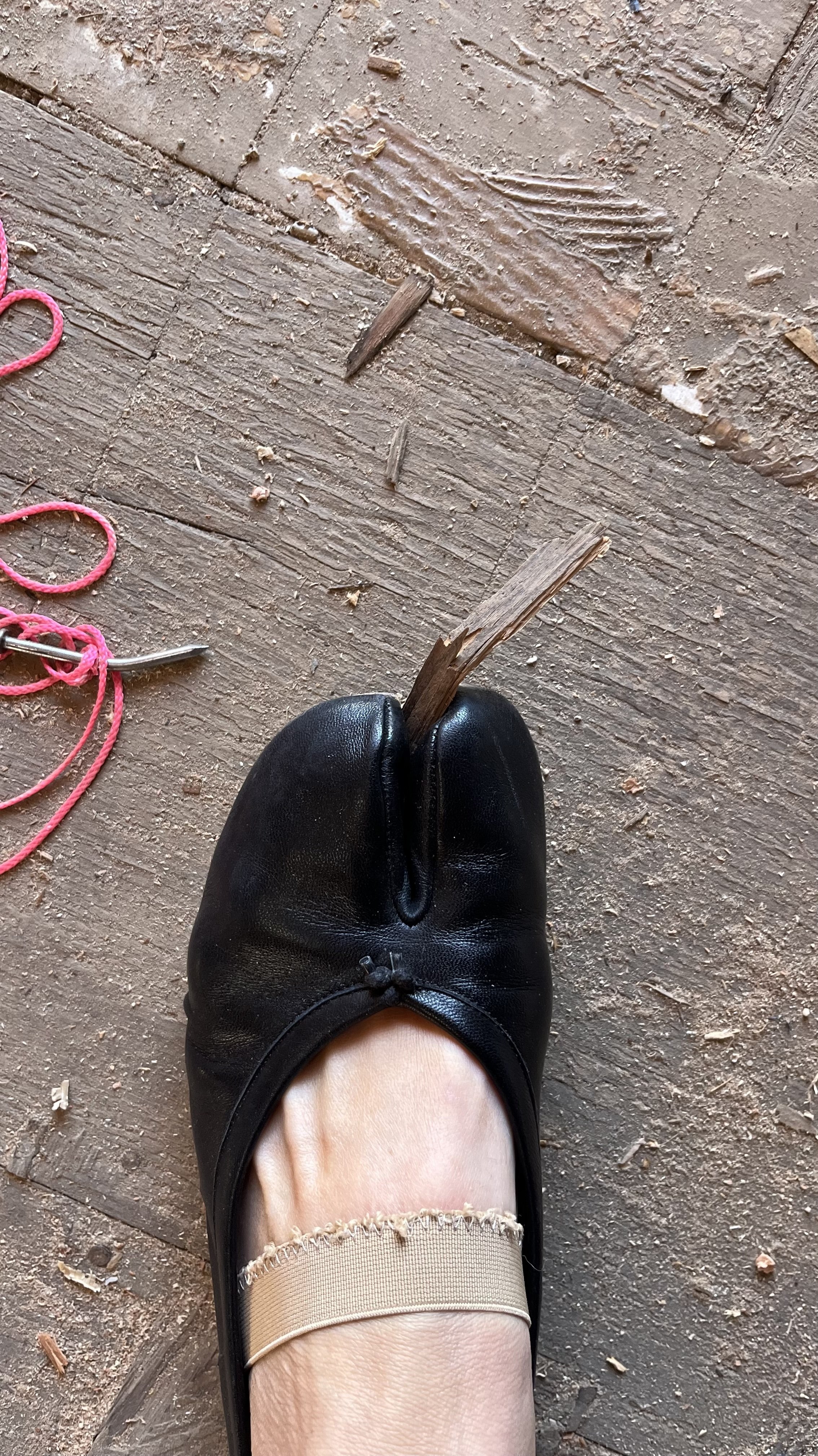 A person wearing a black ballet flat shoe and beige fishnet stocking stands on a wooden floor with debris.