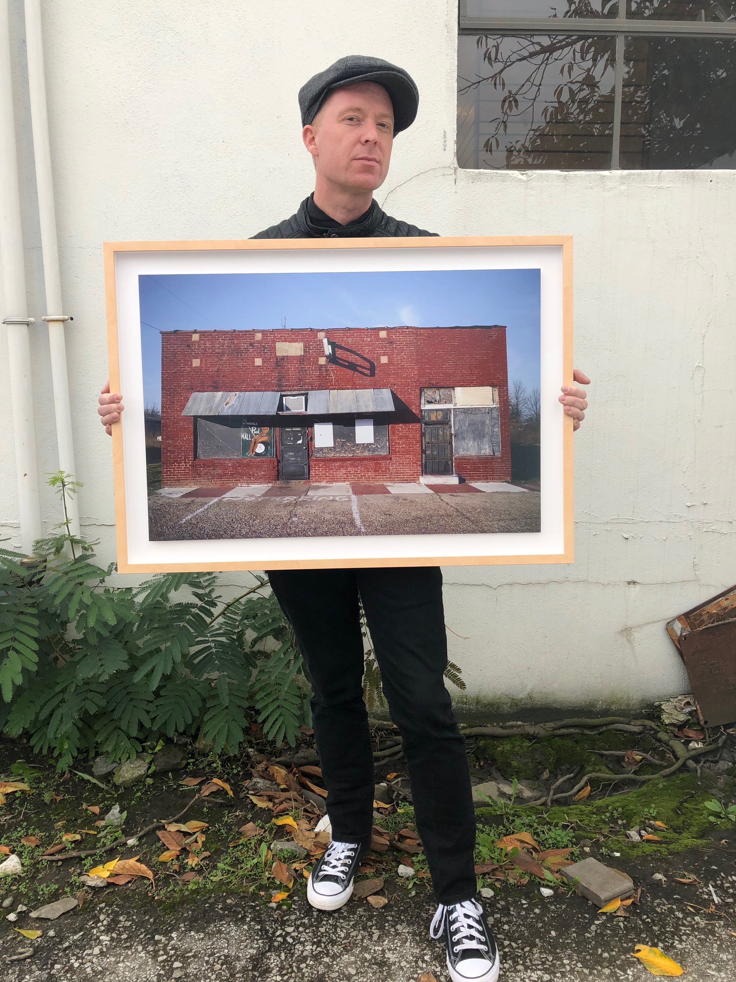 A person stands outdoors holding a framed picture of an old brick building with a metal roof and boarded-up windows, wearing a black leather jacket, black pants, black sneakers, and a grey cap.