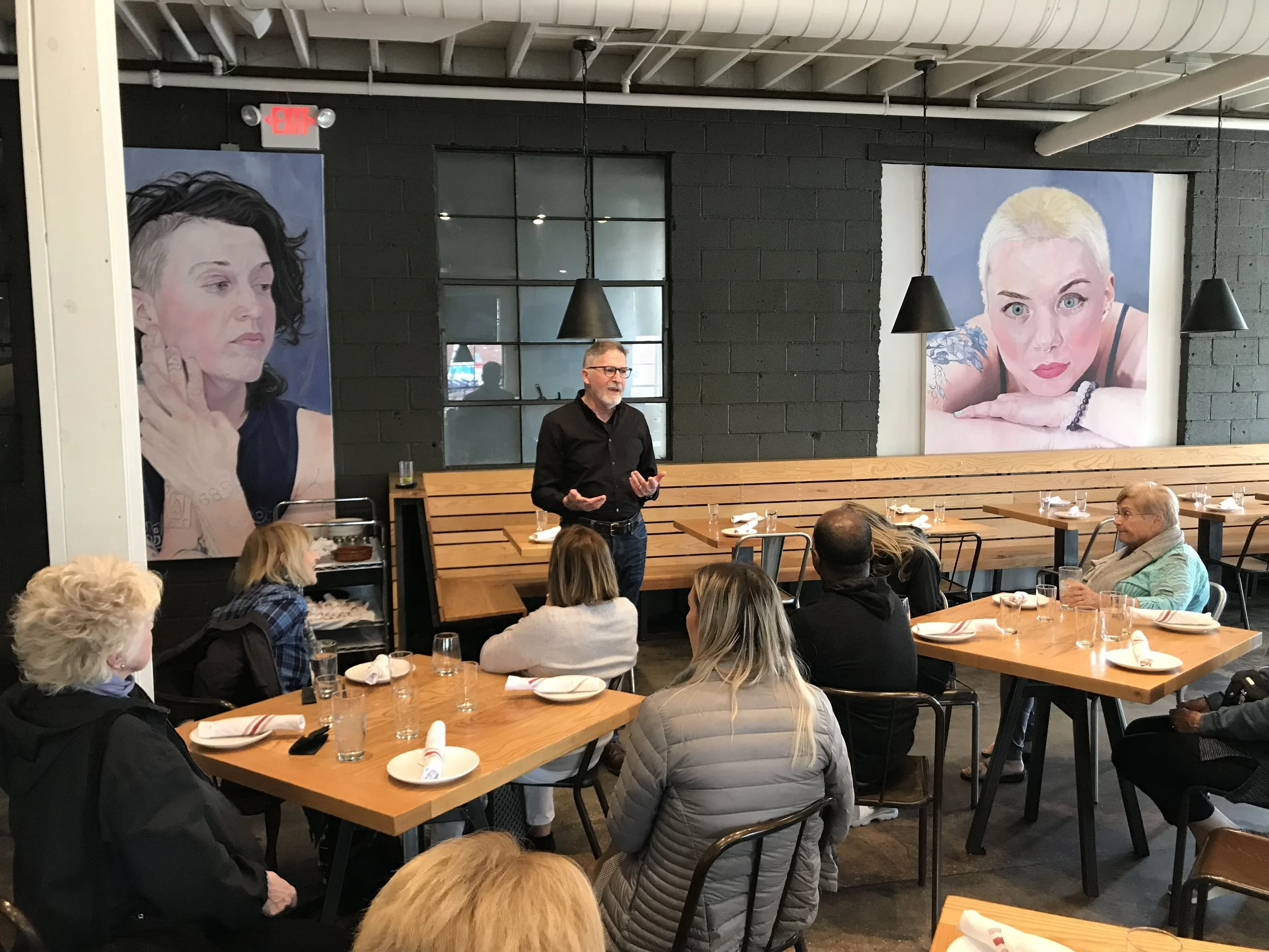 A man in glasses and a black shirt is speaking to a group of people in a restaurant or cafe. There are large portraits of women on the dark brick wall behind him, and the table is set with plates, glasses, and napkins.