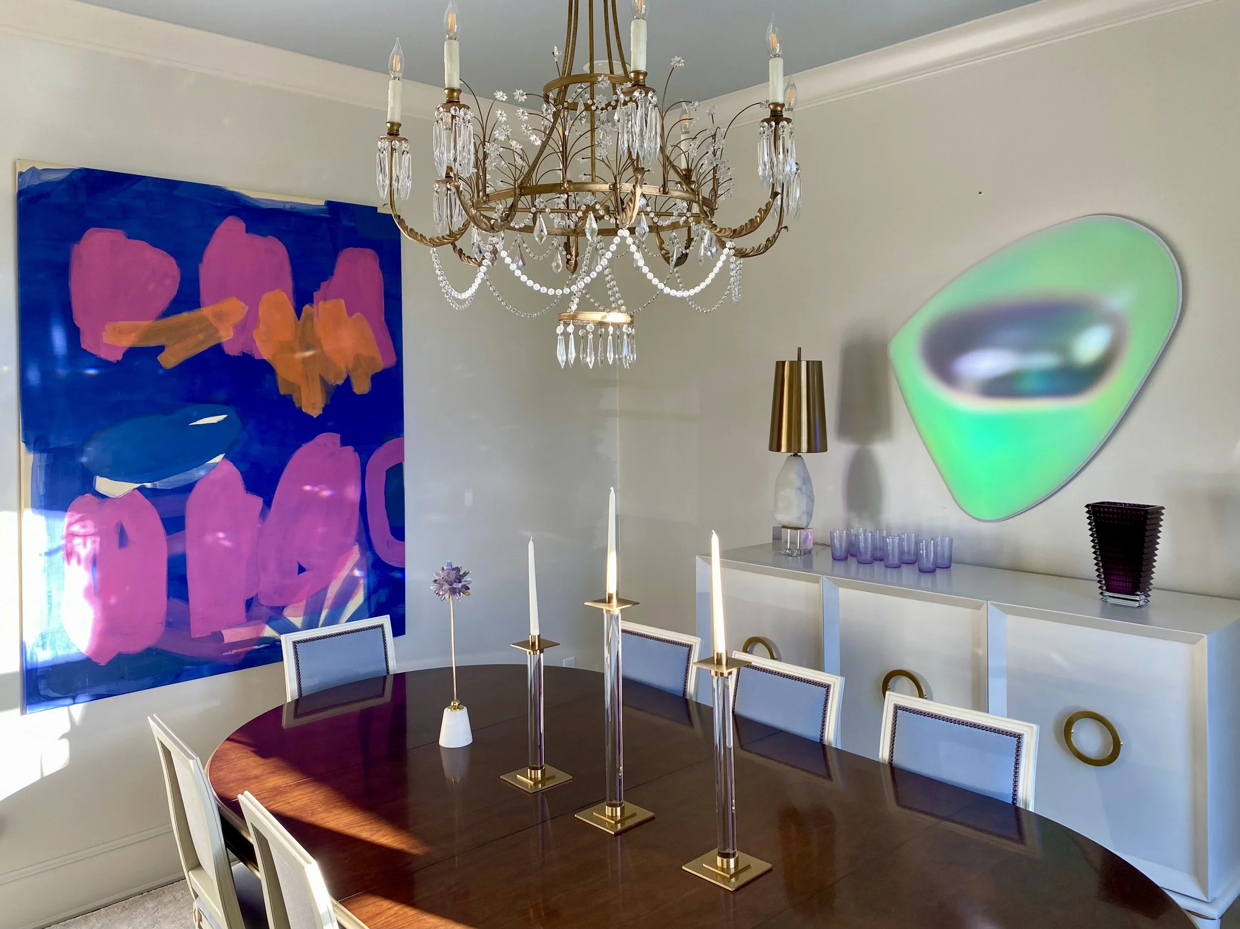 A dining room with a chandelier hanging above a dark wood table surrounded by white chairs with dark cushioned seats. On the wall are two large abstract paintings, one with bold pink and orange shapes on a blue background, and the other with a glowing green and black oval shape.