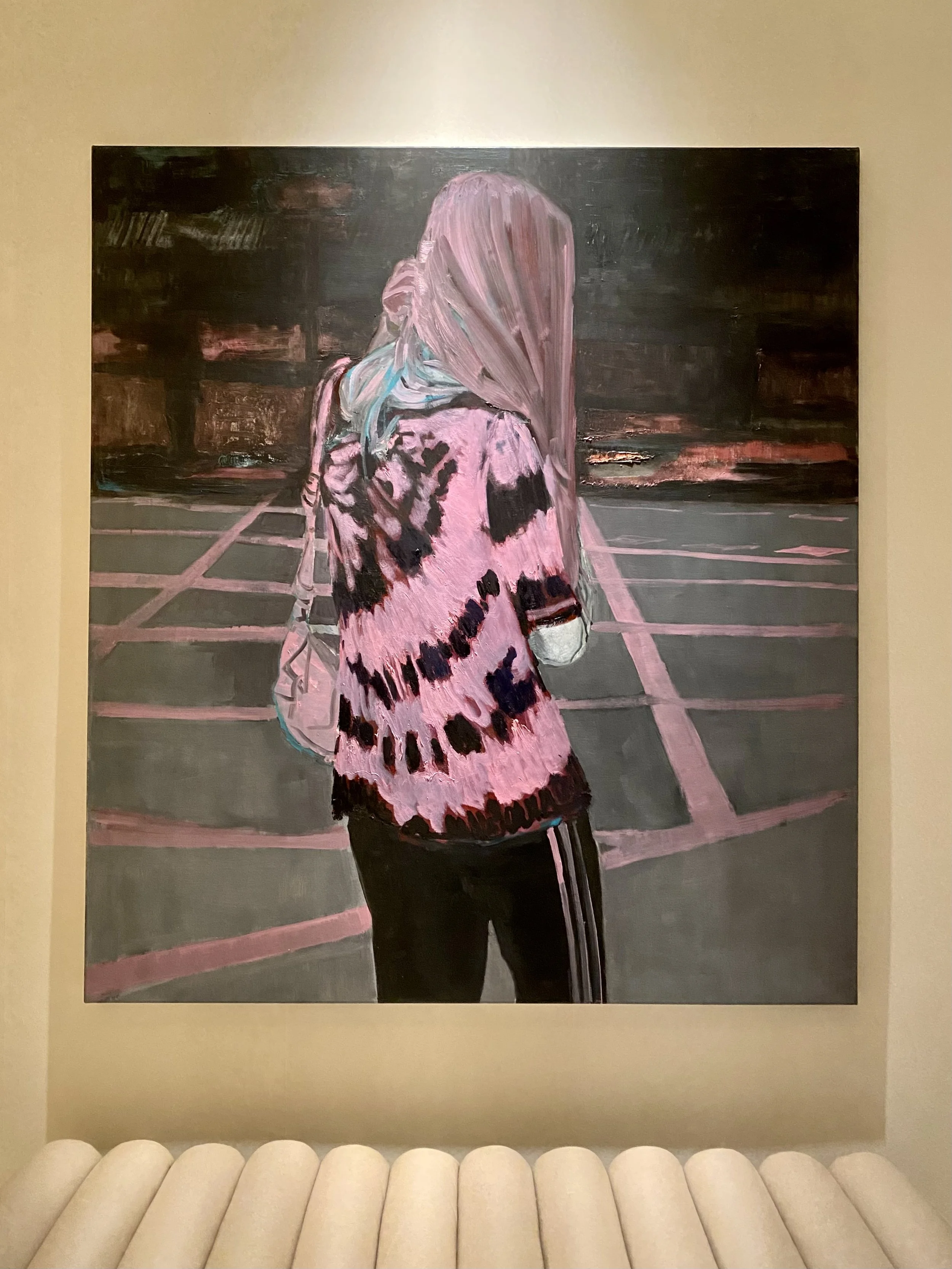 Oil painting of a woman with long hair and a pink and black patterned jacket, standing on an empty parking lot at sunset, with a city skyline in the background.