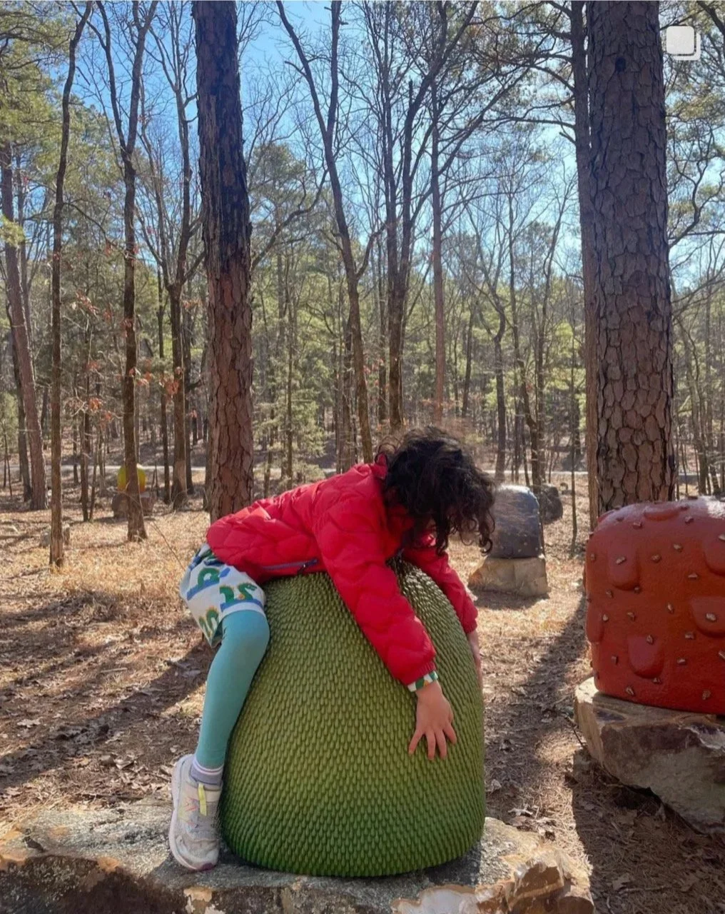 Child climbing on a large, green, textured outdoor sculpture in a wooded area with tall trees and rocks.