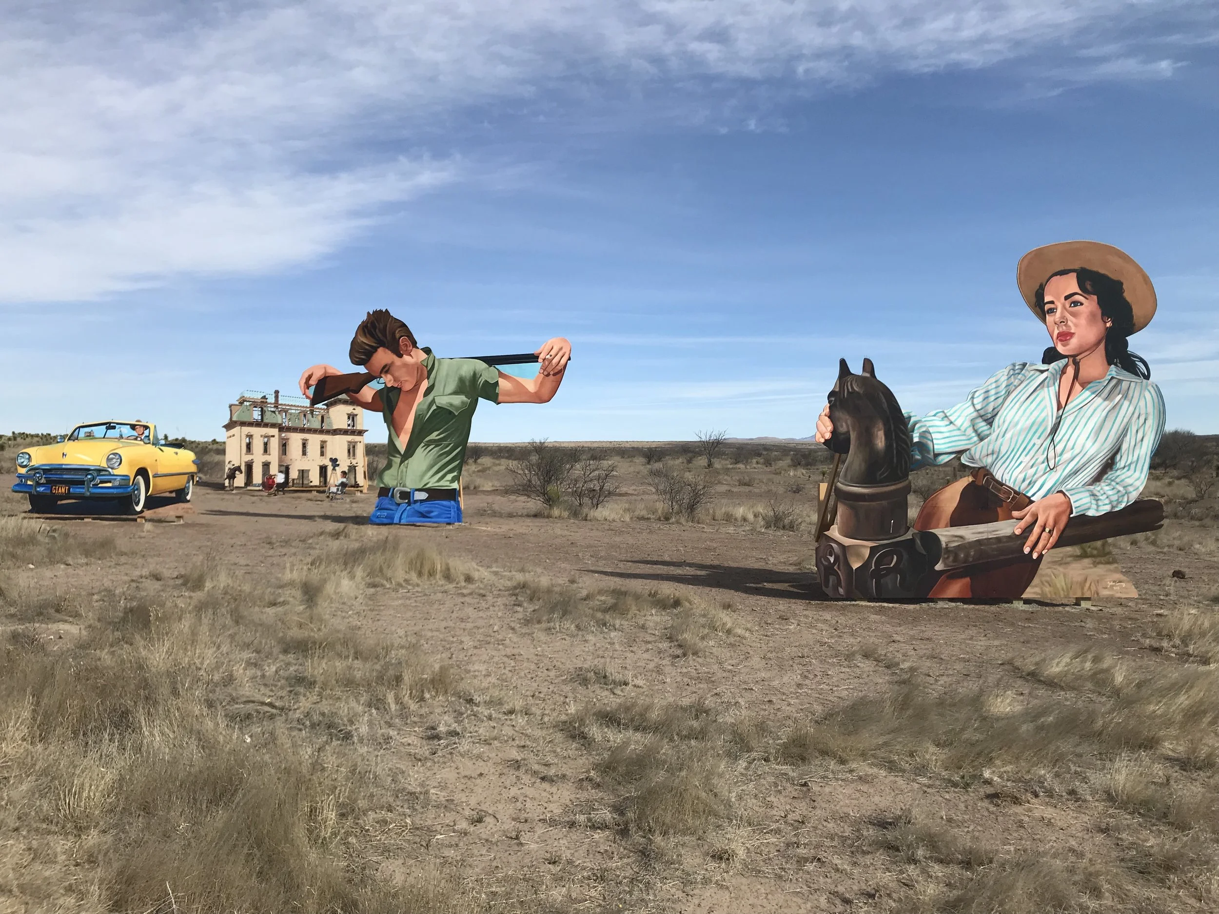 Colorful mural depicting a young man with brown hair and a woman with black hair and a straw hat riding a horse, set against a desert landscape with a clear blue sky and sparse vegetation, with a yellow vintage car and a building in the background.