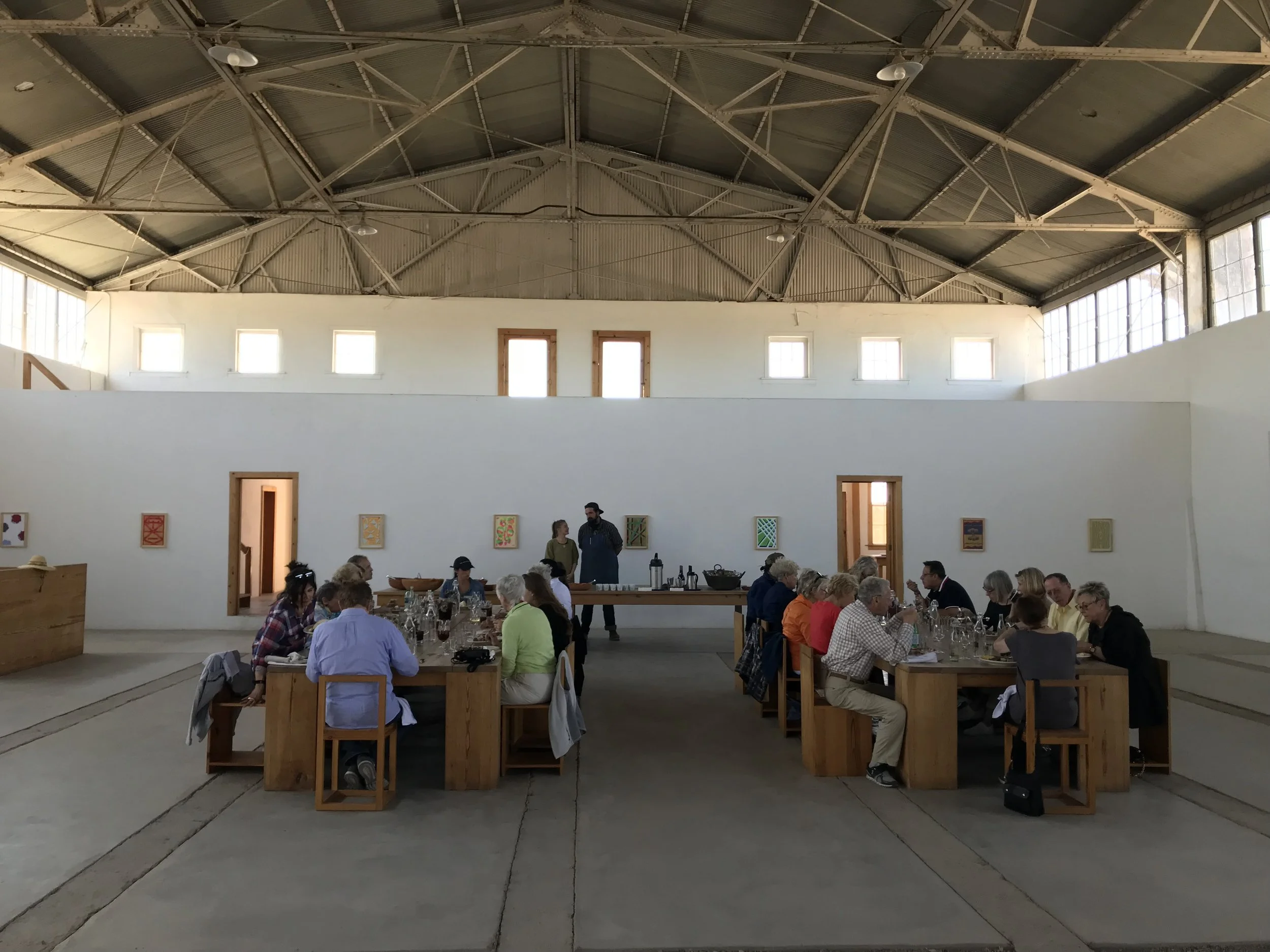 People sitting at tables inside a large, industrial-style building with high ceilings, windows, and exposed metal beams, likely attending a social gathering or event.
