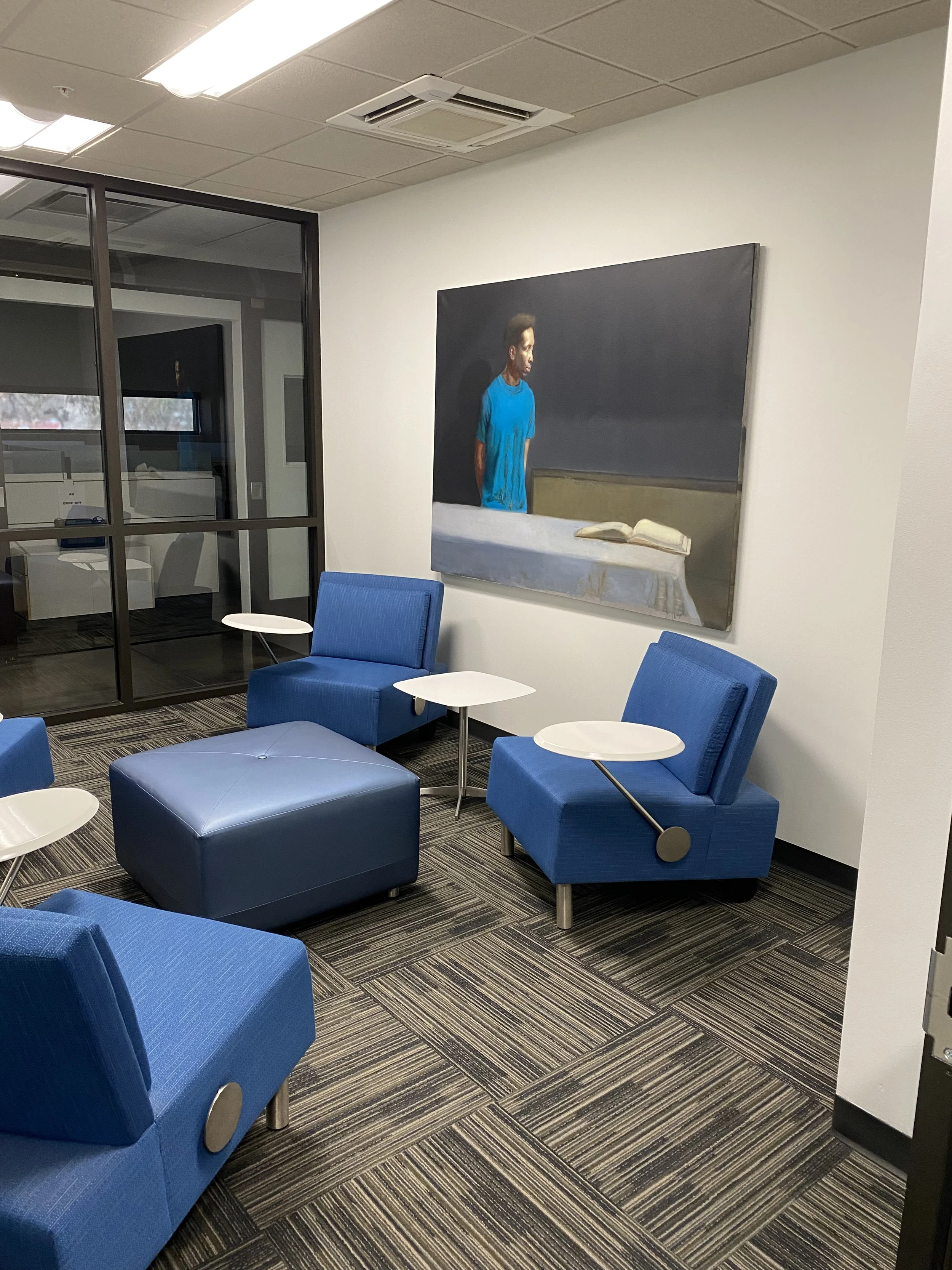 An office waiting area with blue chairs, white tables, a large painting of a person in a blue shirt reading a book on a black background, and a glass partition.