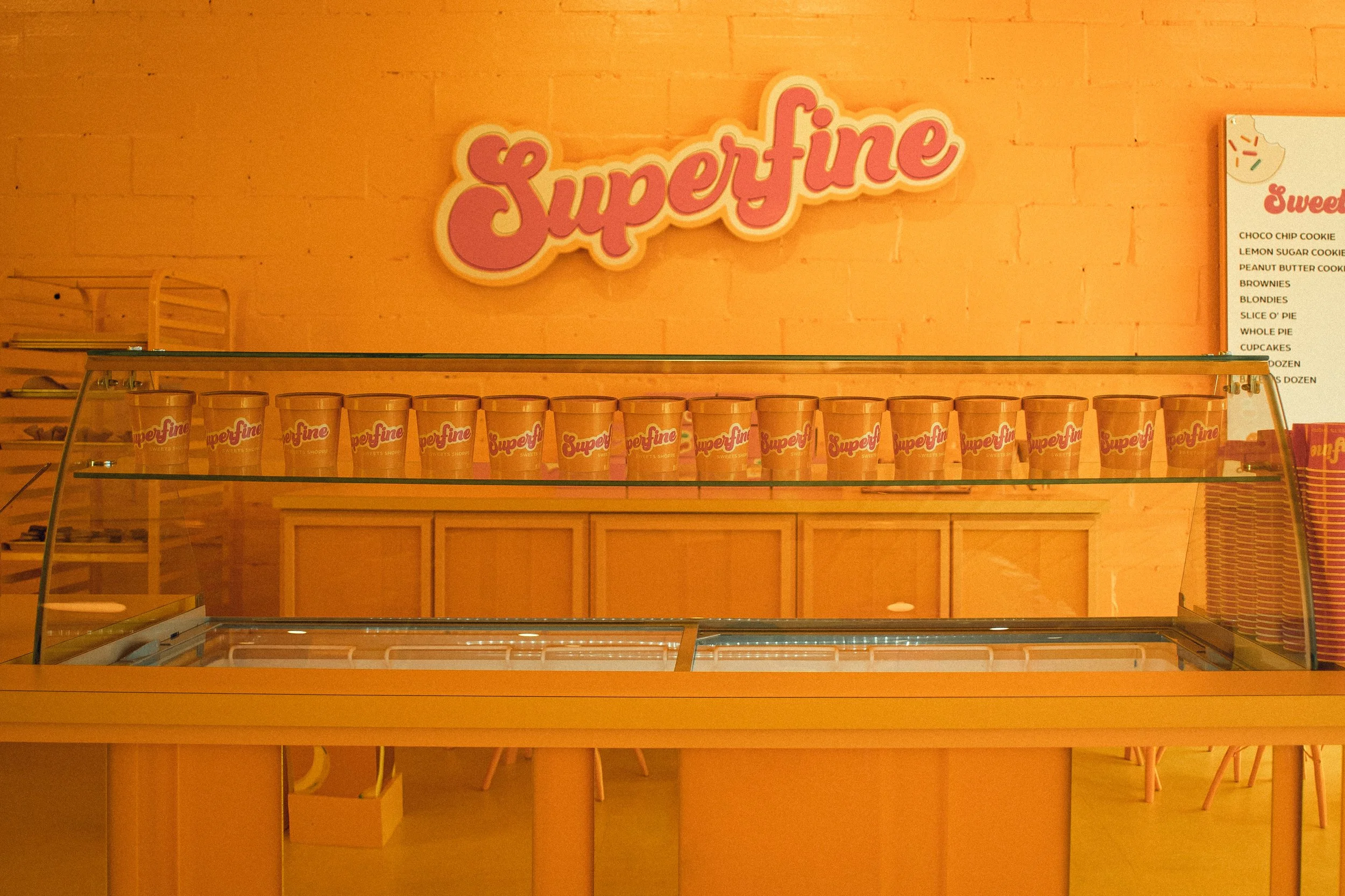 Empty display case with orange cups labeled 'Superfine' in a brightly lit shop or café. Behind the case is an orange brick wall with a pink and white "Superfine" sign. To the right, there is a menu listing various sweet treats.