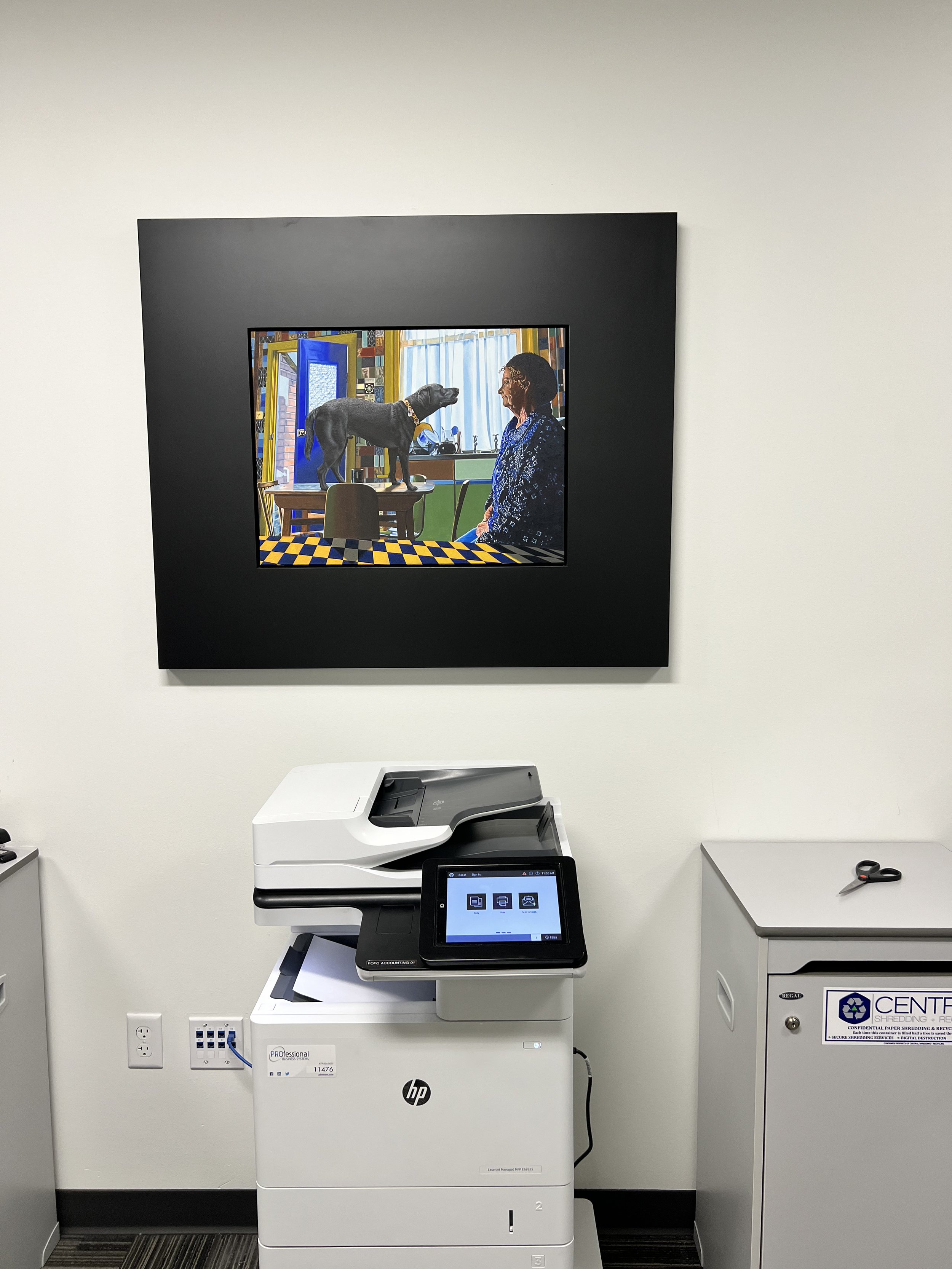 Office printing area with HP multi-function printer and a digital copier in front, a large framed artwork on the wall above depicting a woman and a dog in a colorful kitchen scene, scissors on a small side table, and a white cabinet with recycling and shredding services label.
