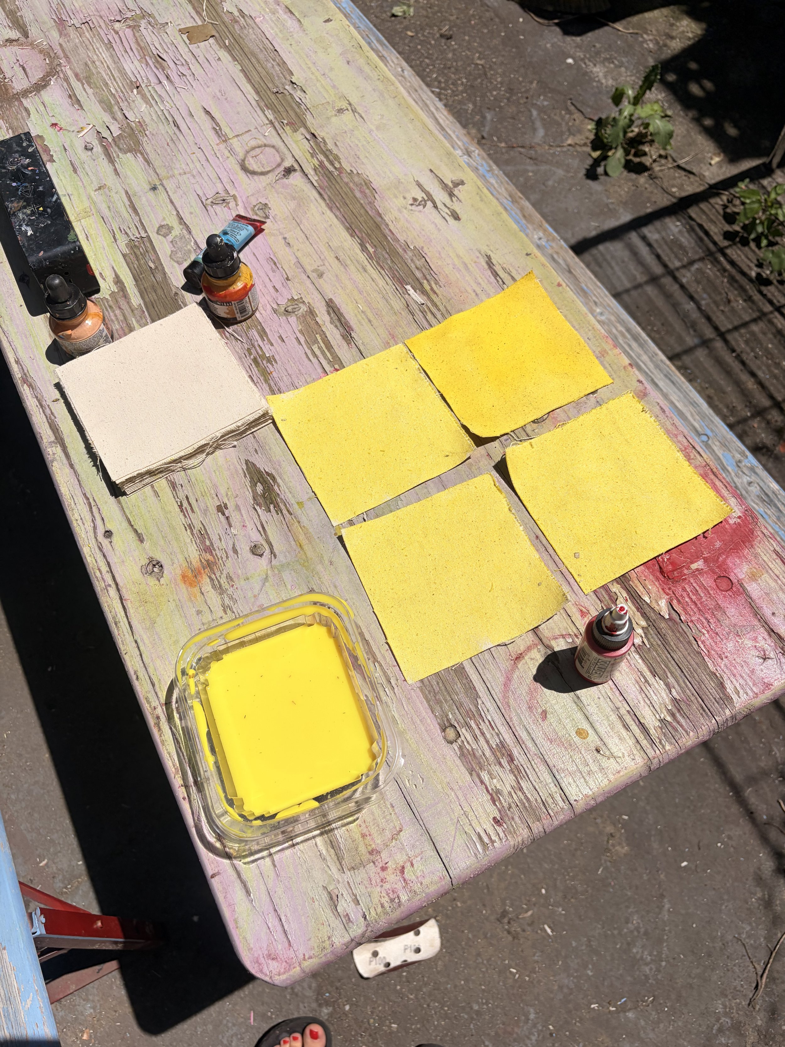 A weathered wooden outdoor table with peeling pink and yellow paint, holding four yellow paint swatches, small bottles of paint, a paintbrush, and a plastic container of yellow paint, with sunlight hitting the surface.