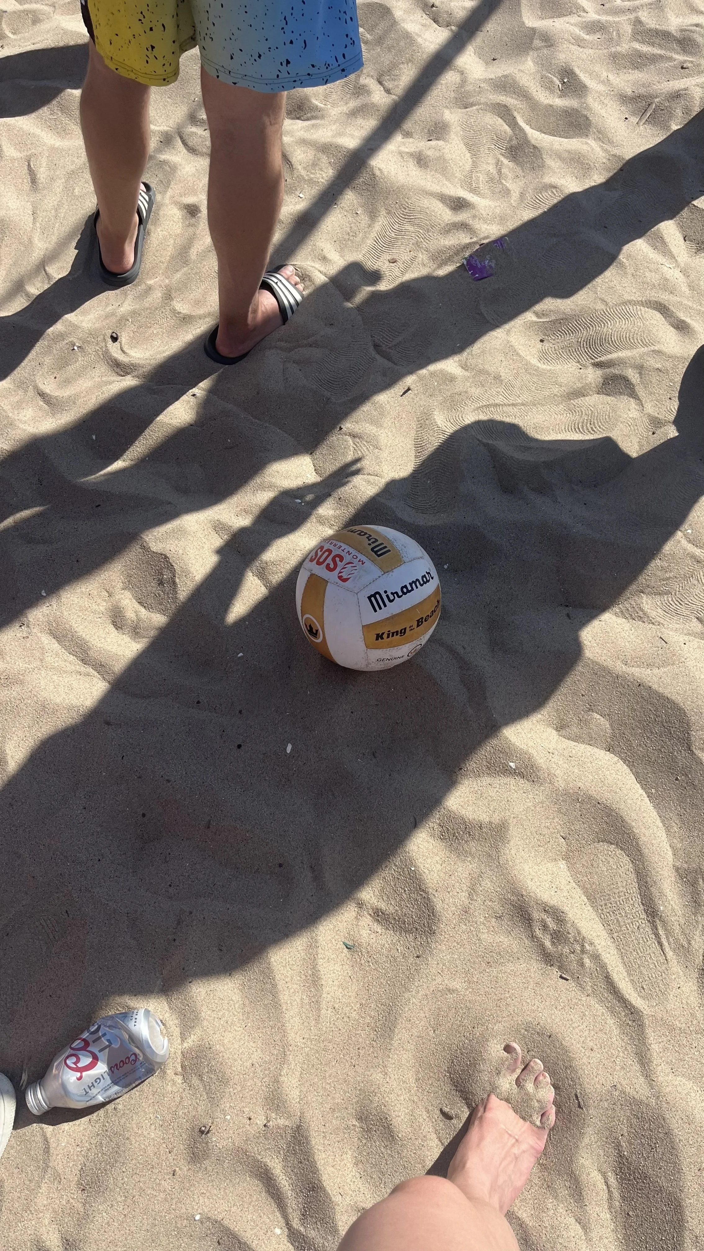 The image shows a sandy beach with a volleyball on the sand, a person’s legs with shorts and sandals, and a foot in the foreground. There are also some discarded bottles and trash in the sand.