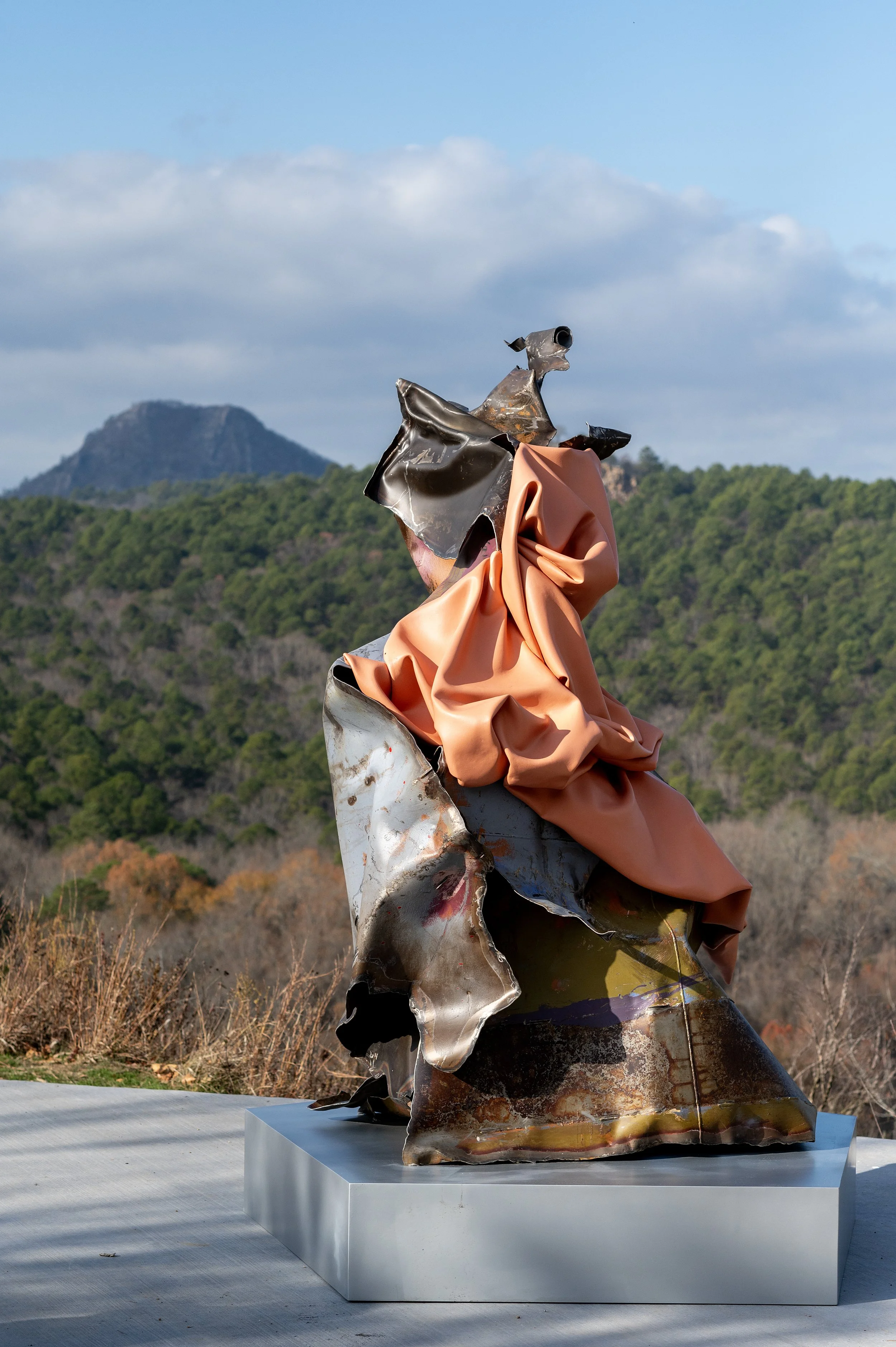 Sculpture art made of metal with rust and paint, featuring a figure with a flowing peach-colored garment overlook mountain landscape with green hills and blue sky with clouds.