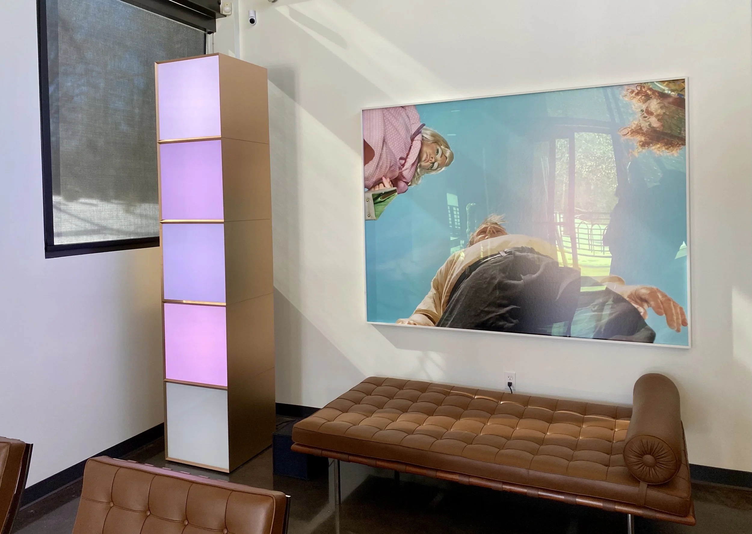 Interior of a modern office or waiting area with a leather bench, a large window reflecting trees outside, and a lighted display or divider with purple lighting.