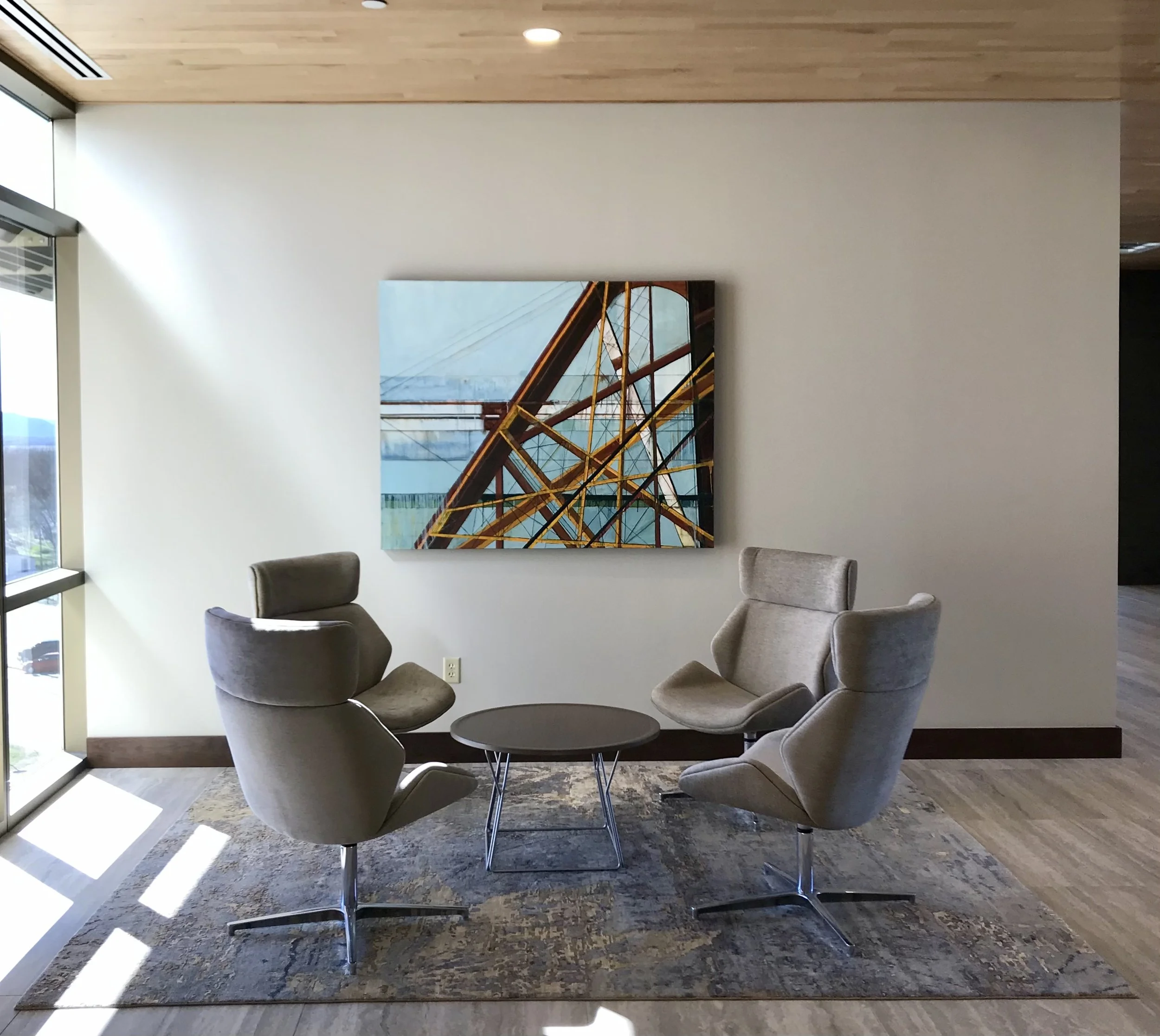 A modern living room with a white wall, a colorful abstract painting featuring a bridge, three gray chairs around a glass table, a window allowing sunlight, and a patterned rug on a wooden floor.