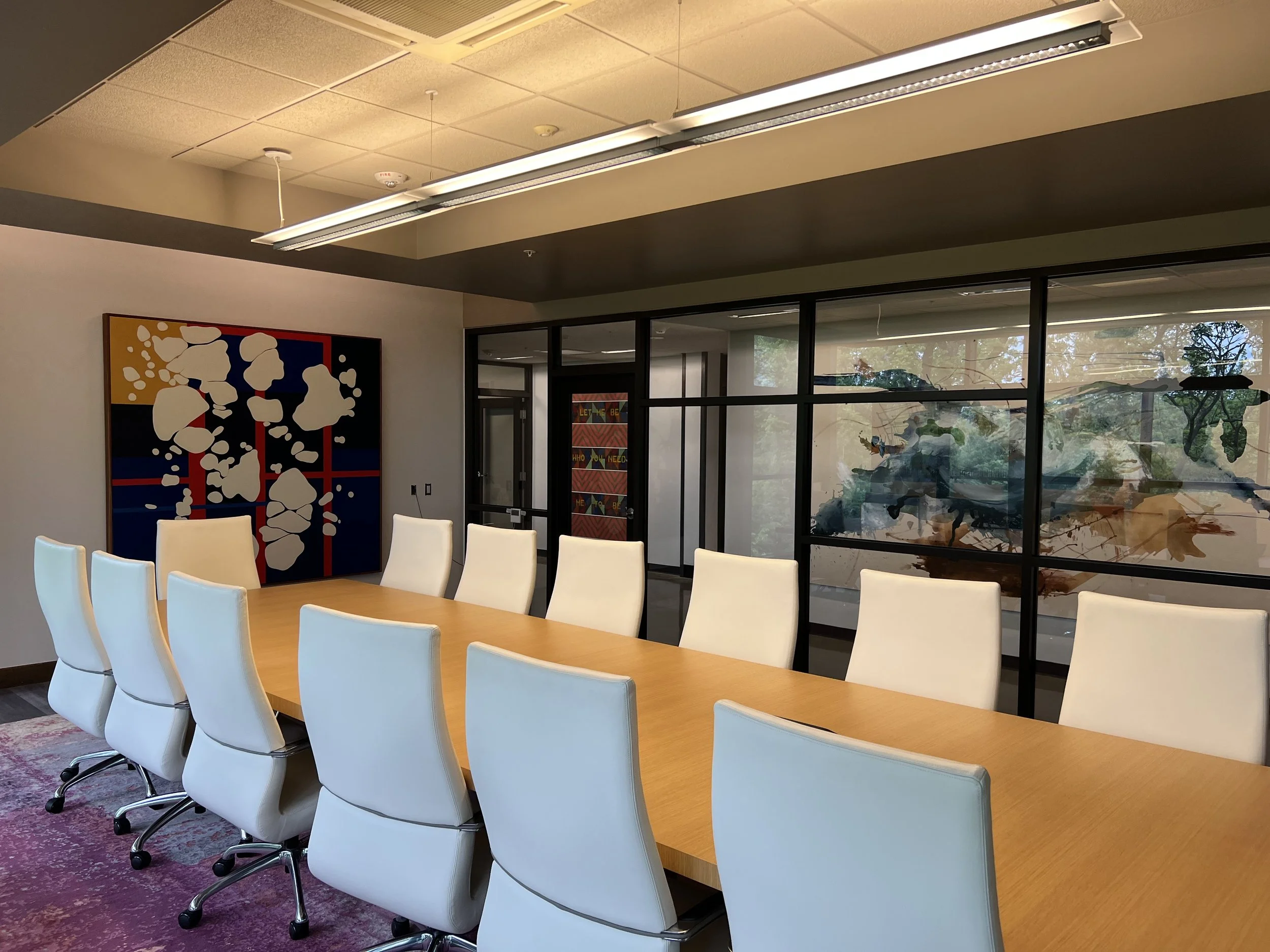 A conference room with a long wooden table surrounded by white chairs, a large colorful abstract painting on the wall, and glass windows showing trees outside. The ceiling has fluorescent lighting.