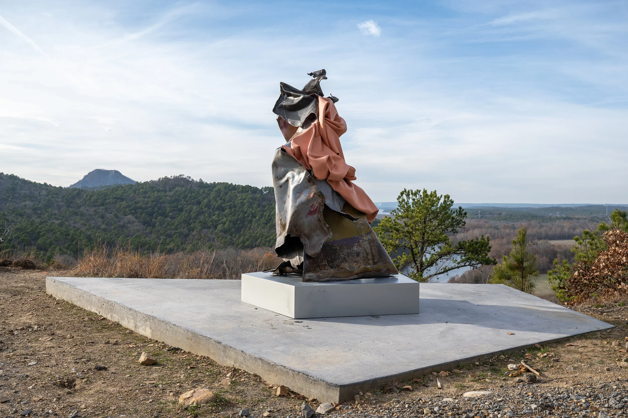 Outdoor sculpture made of metal and fabric on a white pedestal, with a landscape of hills and trees in the background under a blue sky.