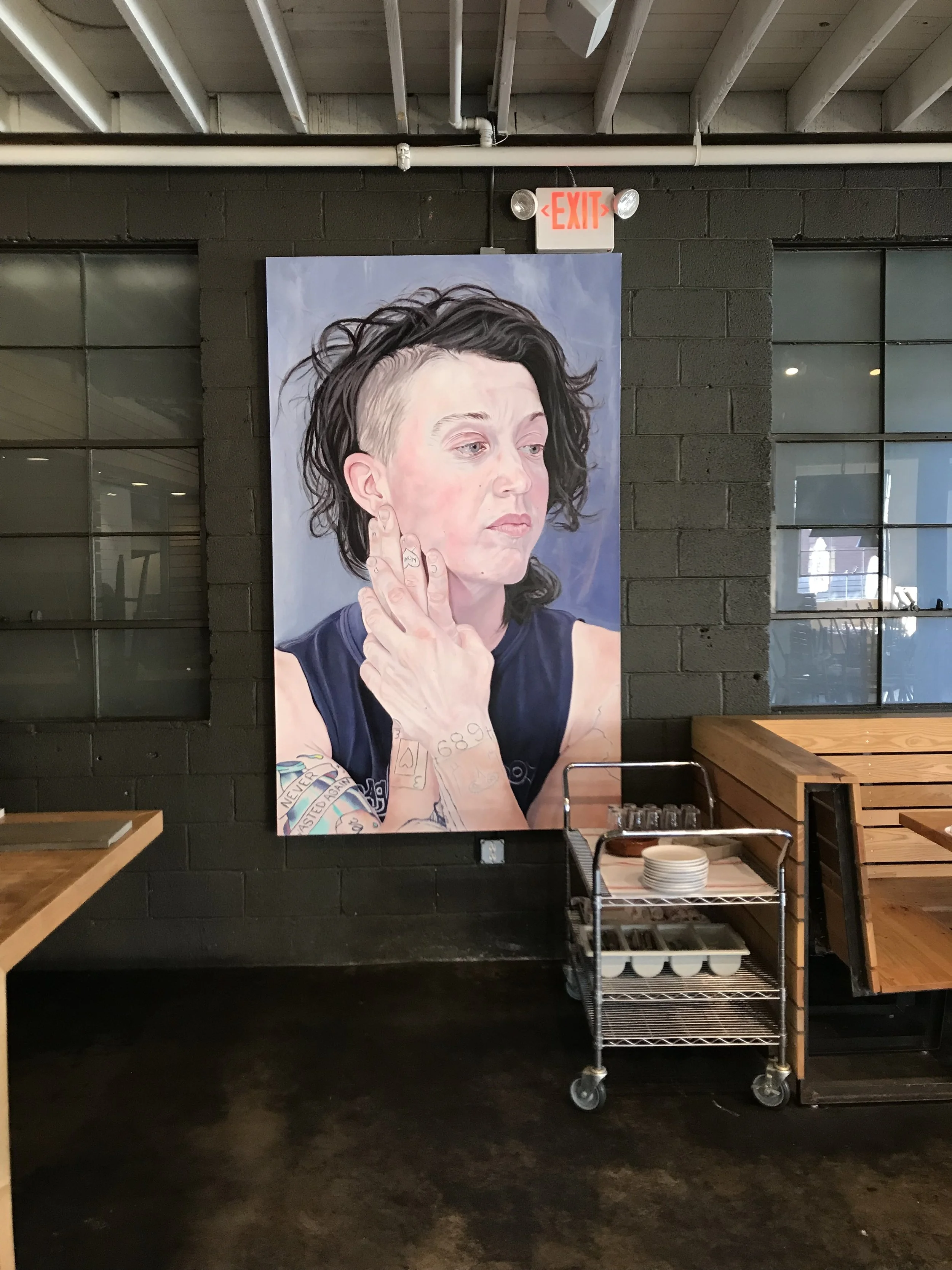 Colorful poster of a woman with short hair and tattoos on her arm, holding her hand to her face, on a black brick wall in a restaurant or cafe.