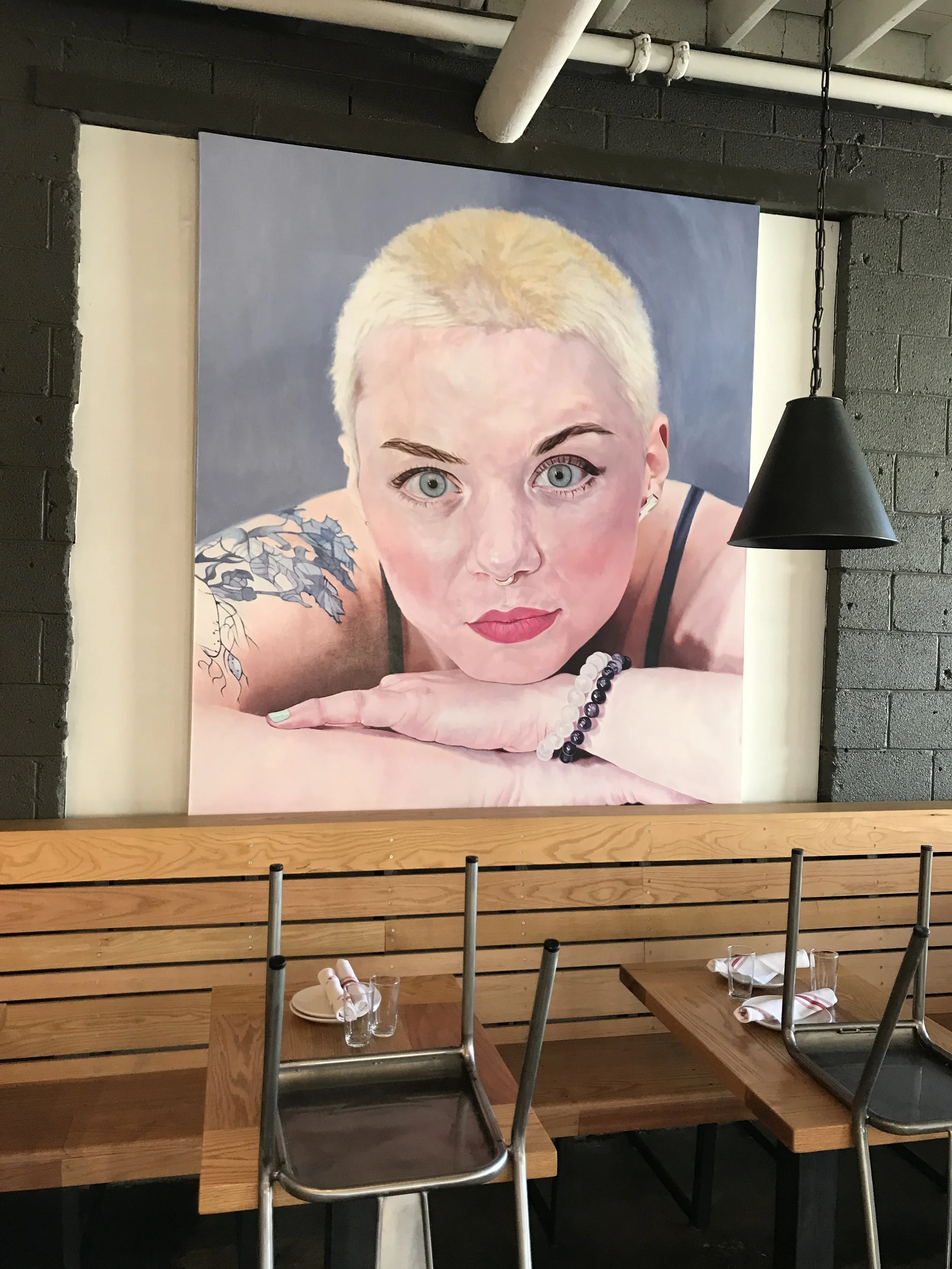 Large portrait painting of a woman with platinum blonde hair, blue eyes, and tattoos on her shoulder, hung on a dark wall in a restaurant with wooden tables and metal chairs.