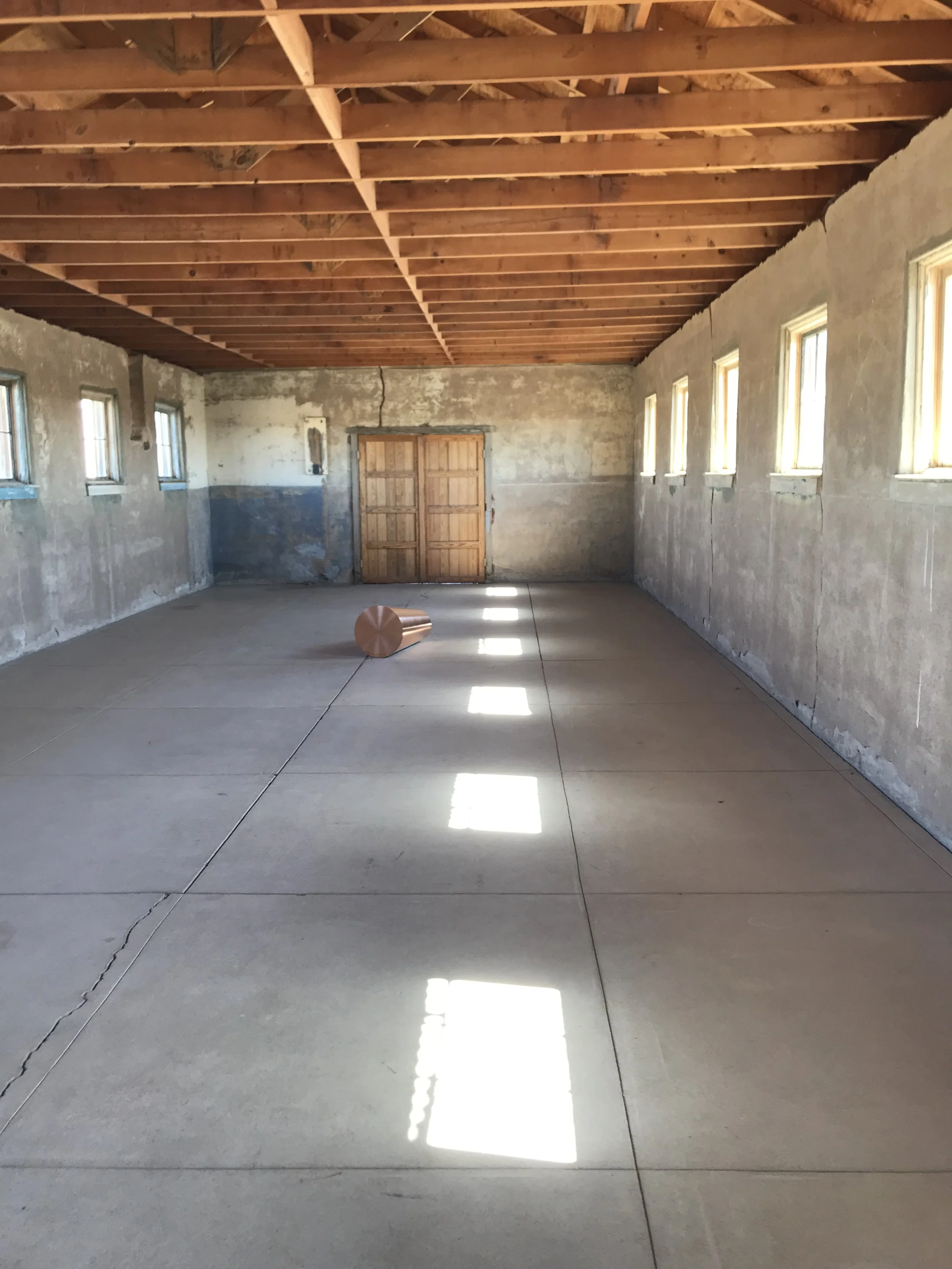 Empty garage or storage space with unfinished walls and a wooden ceiling. Sunlight is coming through multiple small windows on the right, casting rectangular light patterns on the concrete floor. There is an orange object on the floor near the center.