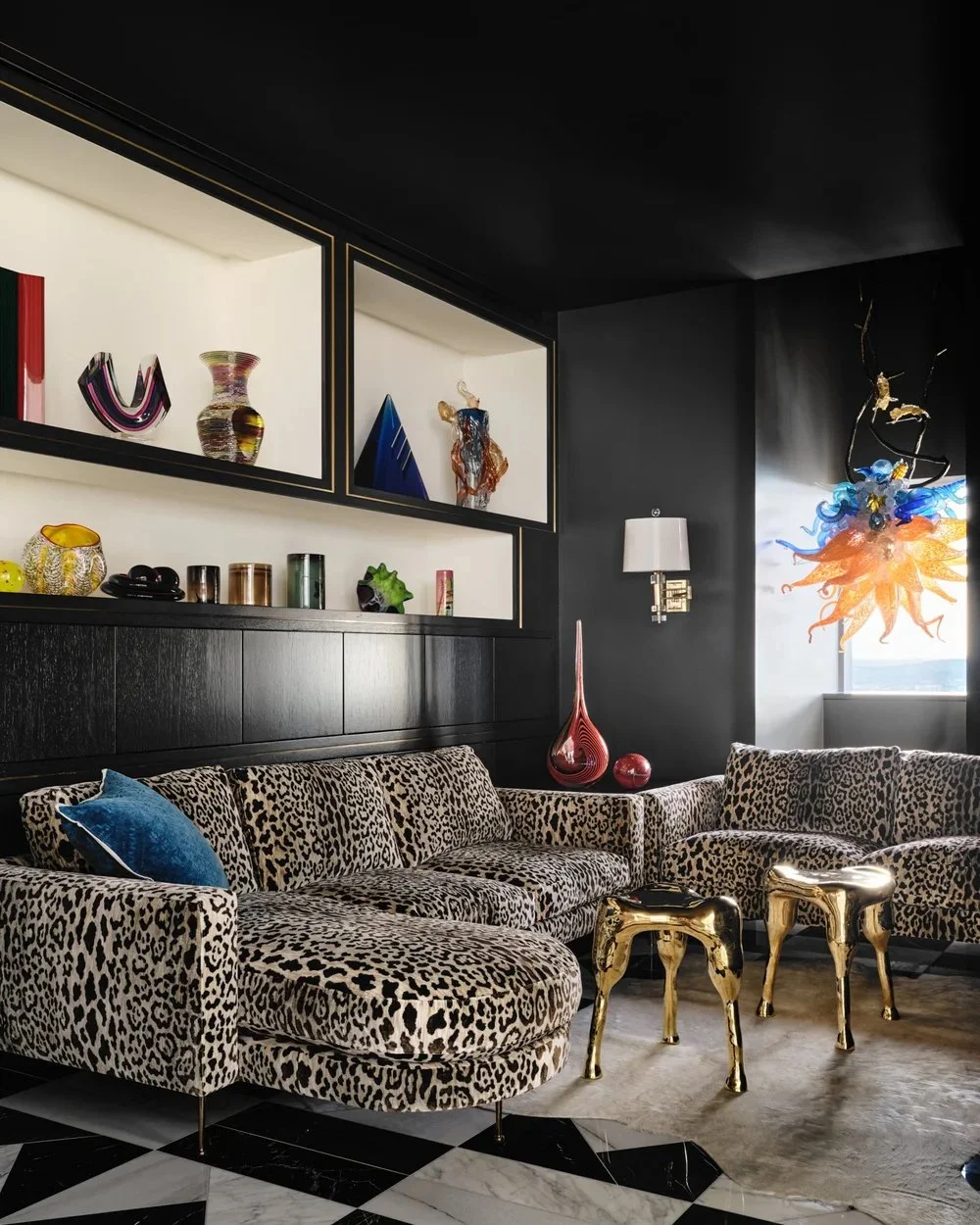 Living room with leopard print sofa, gold metallic side tables, black and white tiled floor, black walls with built-in display shelves holding decorative vases and art pieces, and a colorful glass chandelier near a window.