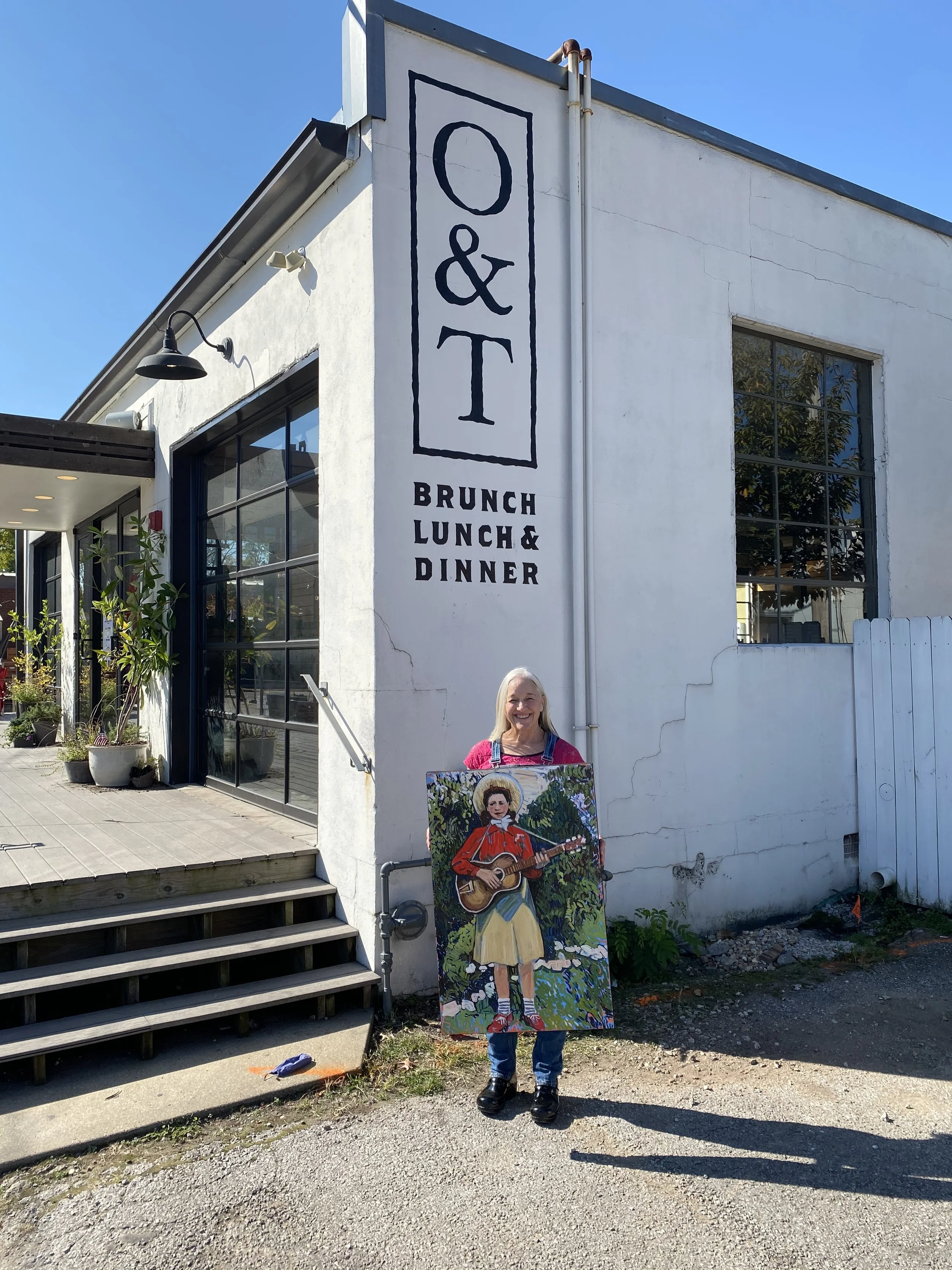 A woman standing outside a white building with large windows, holding a colorful painting of a girl playing guitar. The building has a sign that reads 'O & T Brunch Lunch & Dinner' and there is blue sky above.
