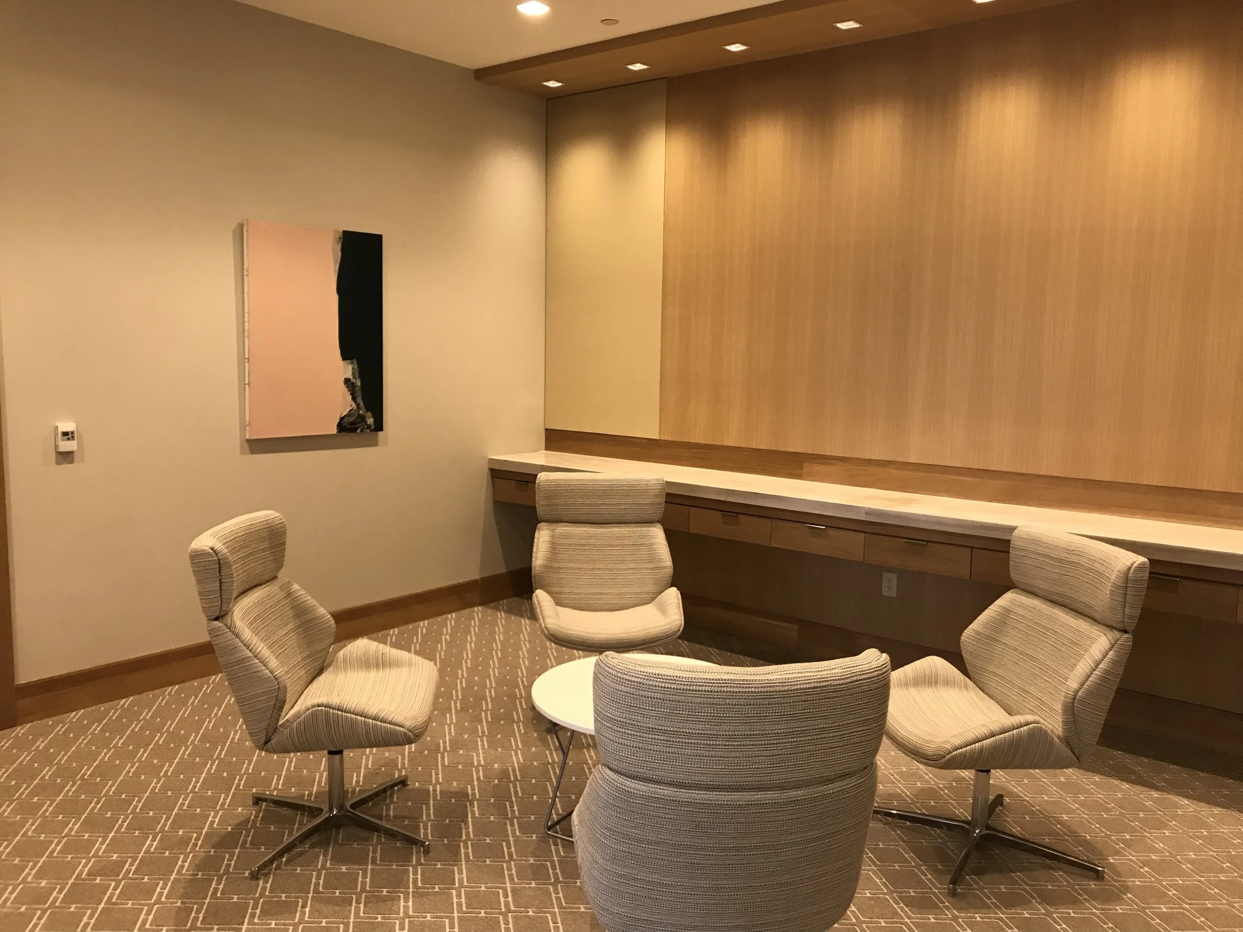 A conference room with beige chairs around a small round table, a wooden wall section with recessed lighting, a beige wall with abstract art, and a beige carpet with a geometric pattern.