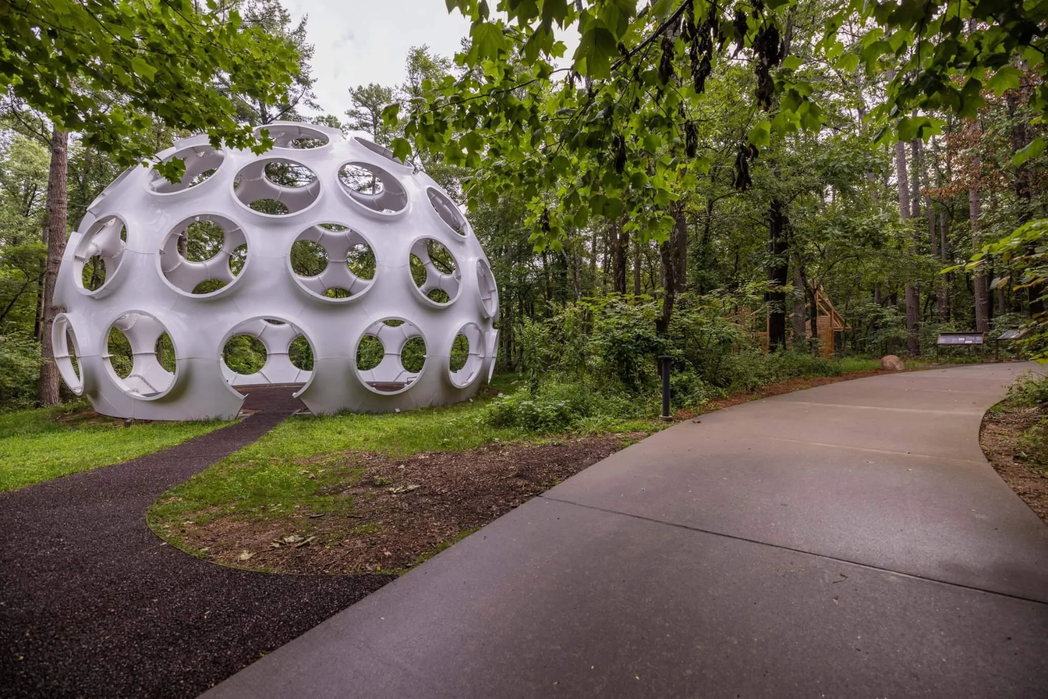 A white geometric structure with circular openings is situated in a wooded area, beside a winding paved pathway.