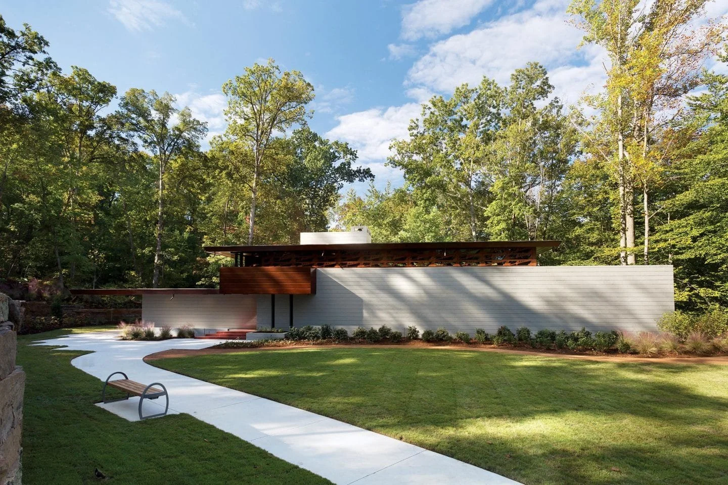 Modern house with flat roof, white and wooden exterior, set in a green landscape with trees, a walkway, and a bench.