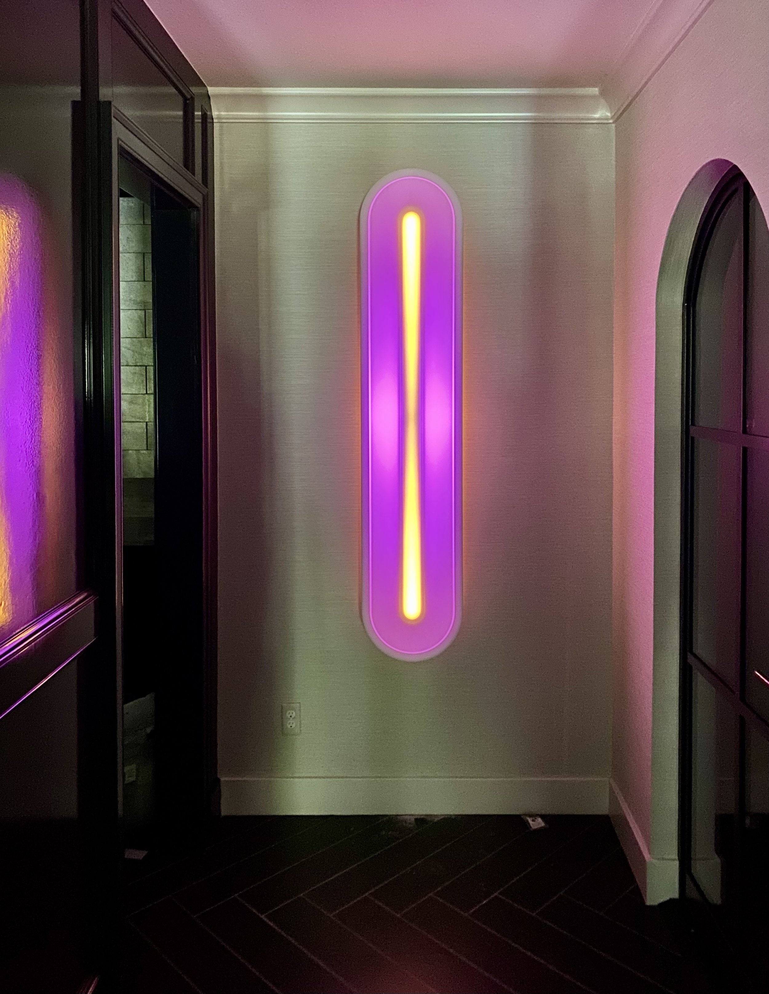 Interior of a room with a vertical rectangular neon sign on the wall in purple, pink, and yellow colors. The room has dark flooring, black-framed glass doors on the left, and a curved window frame on the right. There is a wall socket and an electrical outlet near the floor.